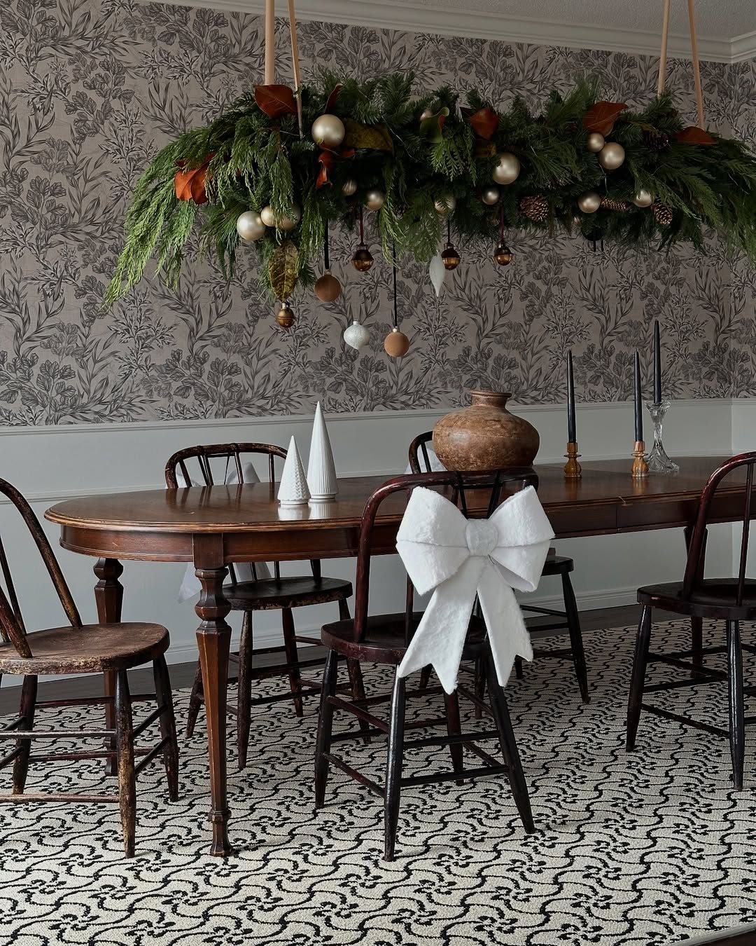 A dining room with a wooden table and chairs, festive greenery and ornaments hanging as Christmas chandelier decor above, and a white bow on one chair. Patterned rug and wallpaper in neutral tones complete the cozy look.