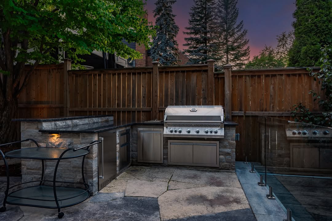 This Rustic outdoor kitchen features a stainless steel grill, stone countertops, and a wooden fence, set in a backyard at dusk with trees and a purple sky—perfect for those seeking inspiring outdoor kitchen ideas.