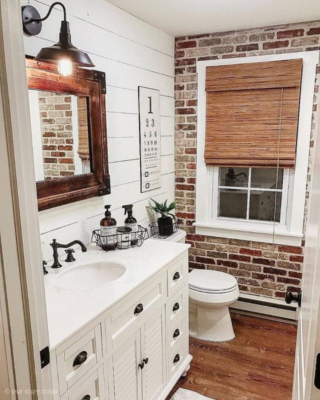 A small shiplap wall bathroom featuring a white vanity, round sink, rectangular mirror, brick accents, toilet, window with a bamboo shade, and an eye chart decoration for a touch of charm.
