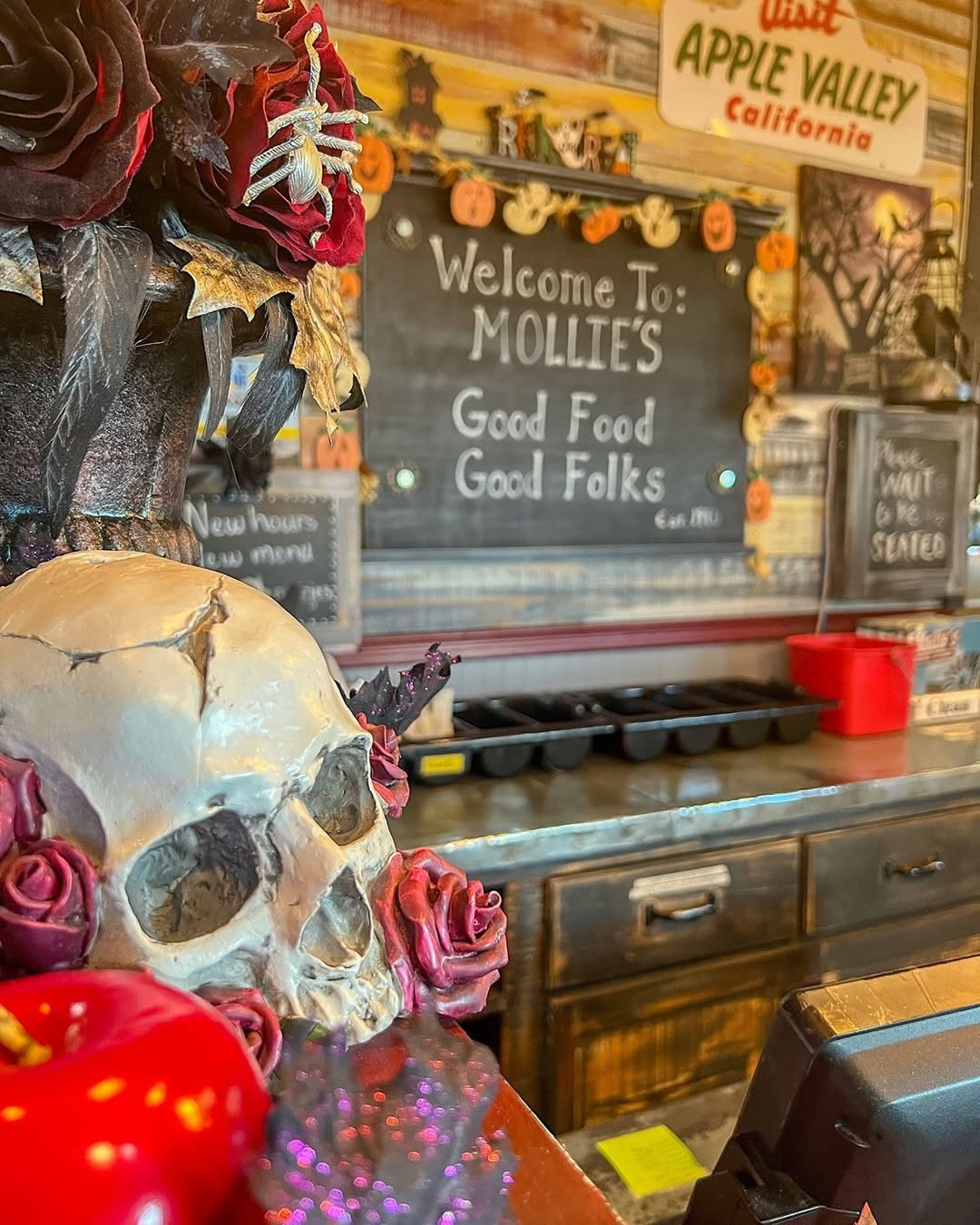 Decorated restaurant counter with Halloween kitchen decor—a faux skull and flowers in the foreground; a chalkboard sign behind reads "Welcome to: Mollie