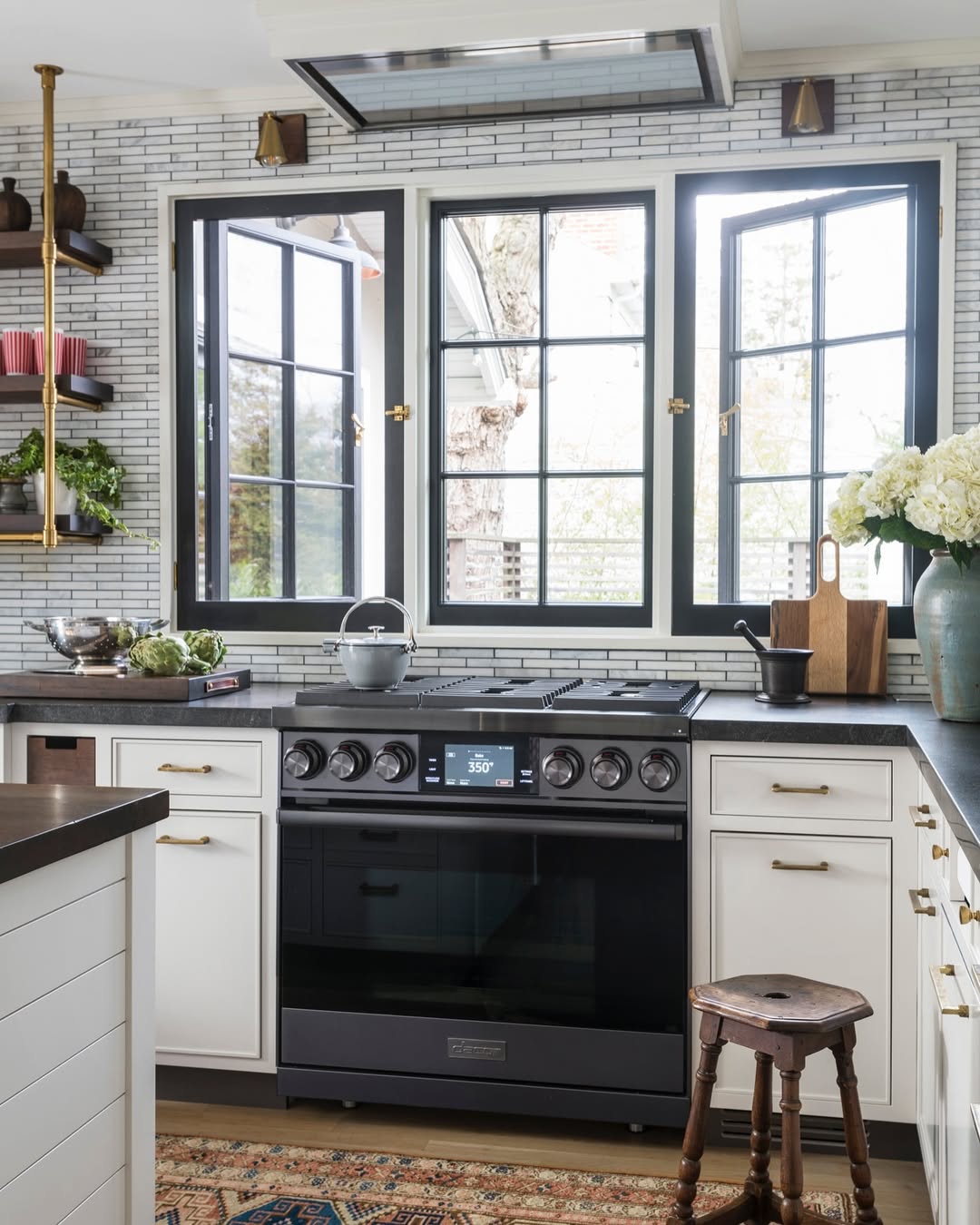 Modern black countertop kitchen with white cabinets, a large window above the stove, open shelving displaying dishes, a kettle, flowers, and a wooden stool in the foreground.