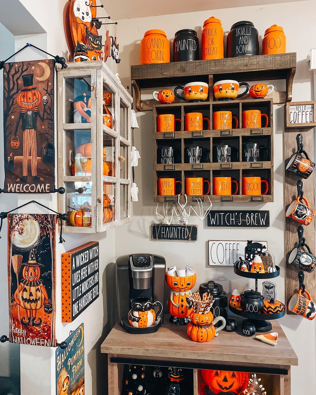 A kitchen corner decorated for Halloween 2025 features orange and black mugs, pumpkin-themed items, wall signs, and festive figurines on shelves and a countertop—perfect for inspiring your own Halloween kitchen decor.
