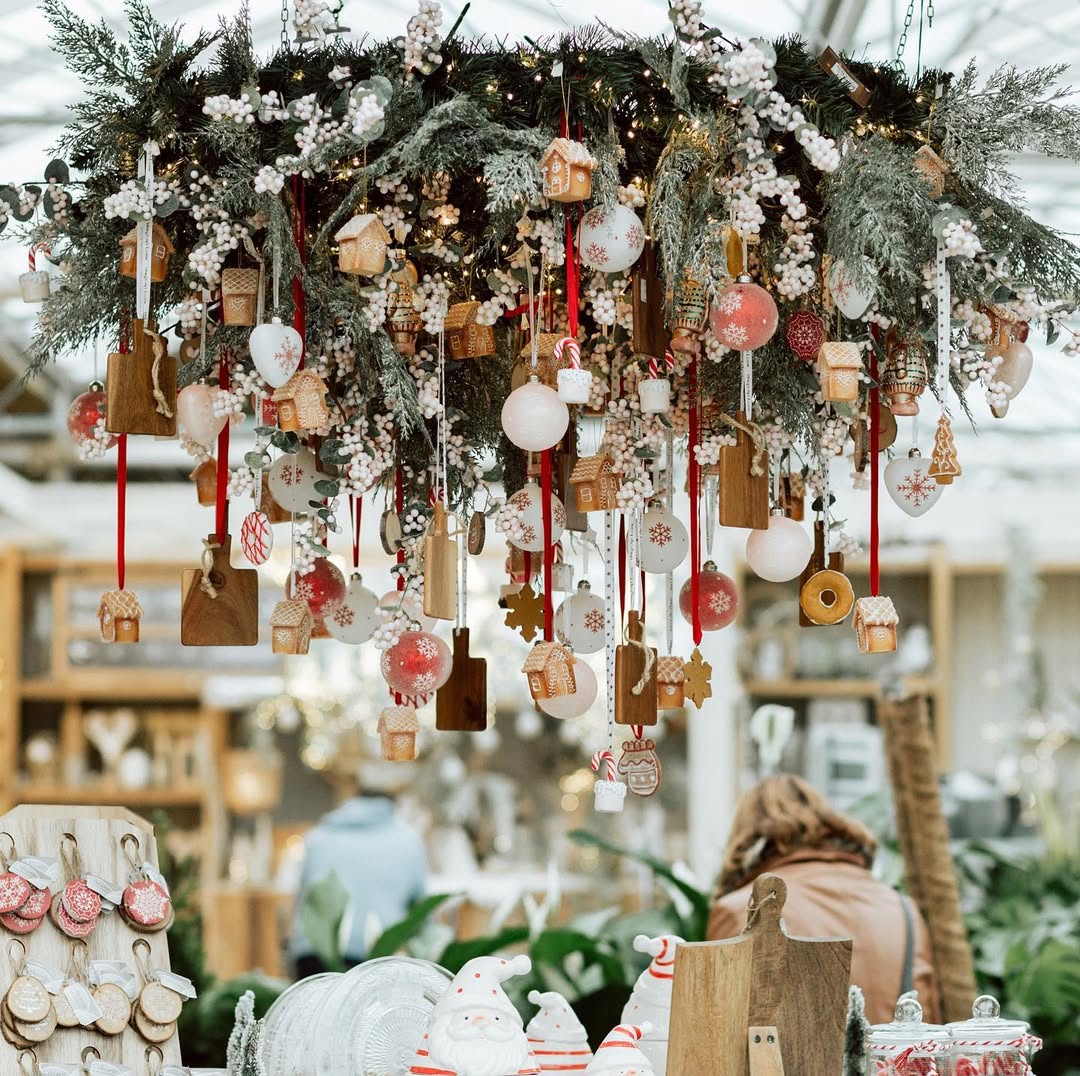 A large hanging arrangement of greenery with ornaments, cookies, and wooden cutting boards creates stunning Christmas chandelier decor above a table in a shop.