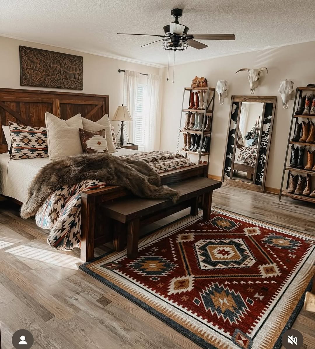 A rustic bedroom featuring ranch house decor: a wooden bed, patterned bedding, a red southwestern rug, a bench, full-length mirror, boot shelves, and animal skulls mounted on cream-colored walls.