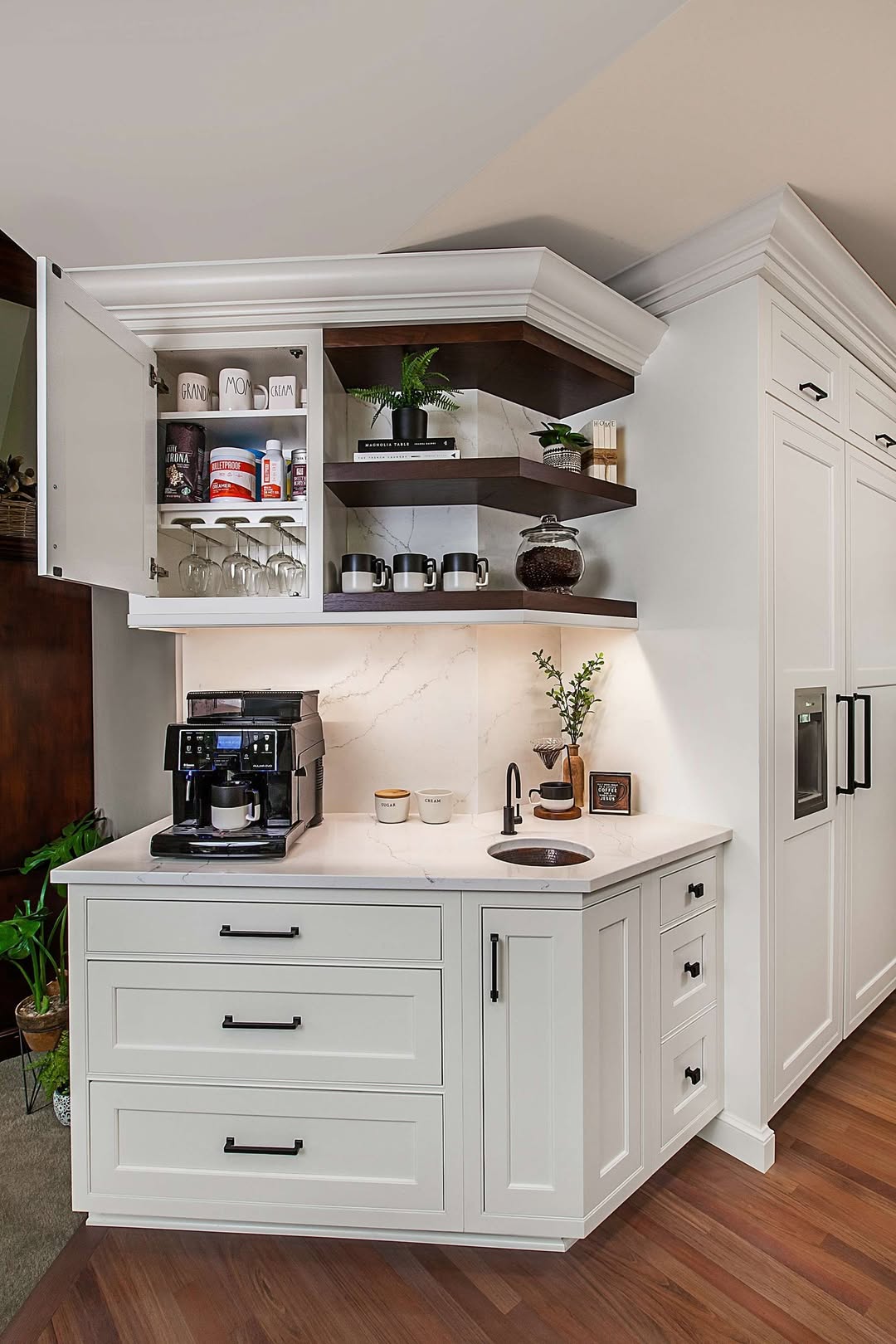 A white corner kitchen cabinet with open shelves, drawers, a coffee maker, cups, decor items, a sink, and under-cabinet lighting on a wood floor—perfect for inspired Kitchen coffee bar ideas without countertop chaos.