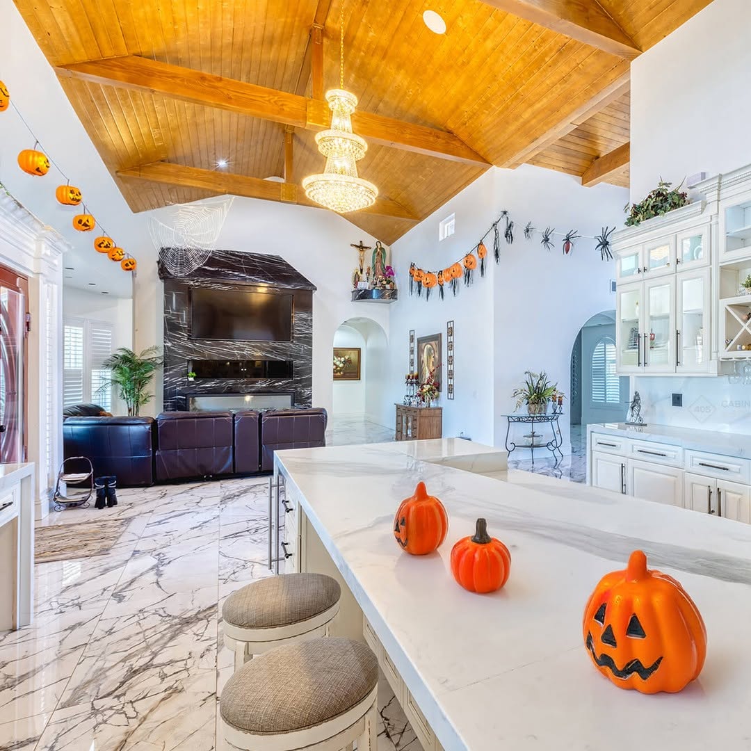 A bright, spacious kitchen and living area decorated with Halloween 2025 pumpkins and festive garlands, featuring marble floors, a wooden vaulted ceiling, and inspiring Halloween decor ideas for your kitchen.