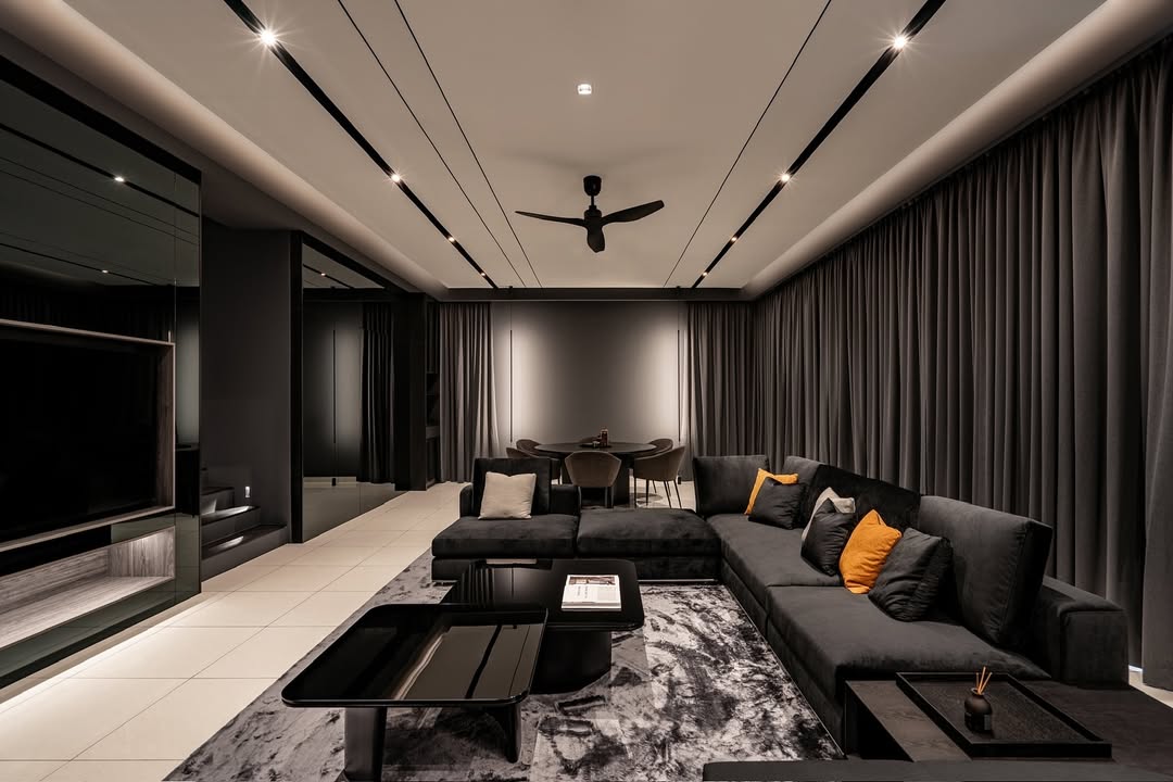 A modern black living room with sleek black sofas, orange accent pillows, a black coffee table, gray rug, and dark curtains. A ceiling fan and dining area can be seen in the background.