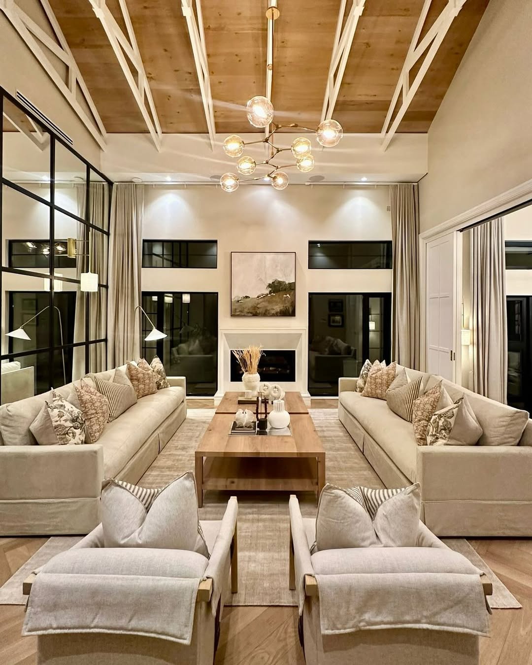 Modern living room with high ceiling, two beige sofas, armchairs, a wooden coffee table, large windows with curtains, and a fireplace with artwork—perfect inspiration for high ceiling living room ideas to make your home feel bigger.