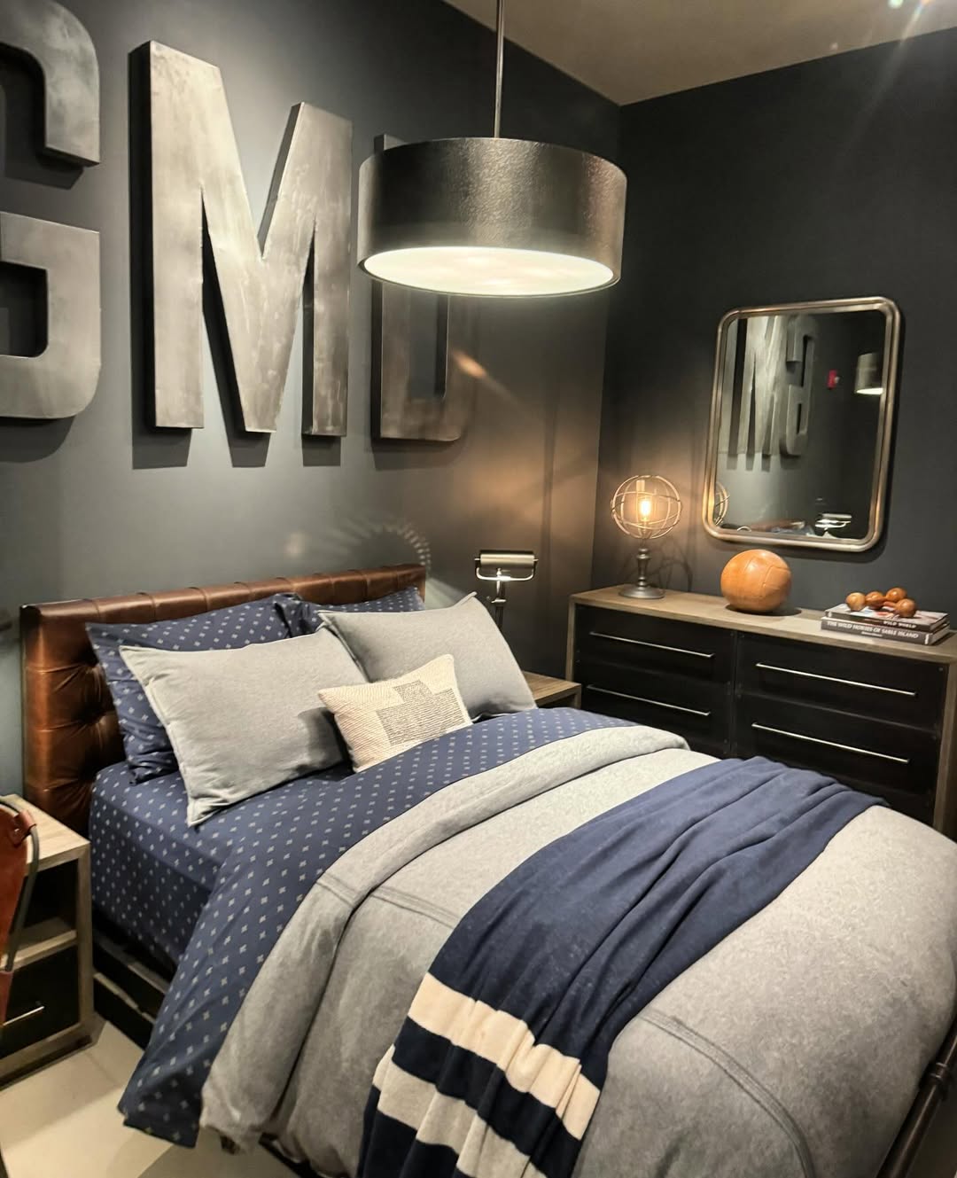 Modern boys bedroom with dark walls, large metallic letters on the wall, a neatly made bed with blue and grey bedding, a round ceiling lamp, matching nightstands with decor, and smart storage solutions for sleep optimization.