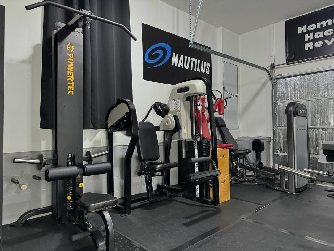 A mini gym at home featuring multiple exercise machines, including Powertec and Nautilus equipment, neatly arranged against the wall in a spacious garage.
