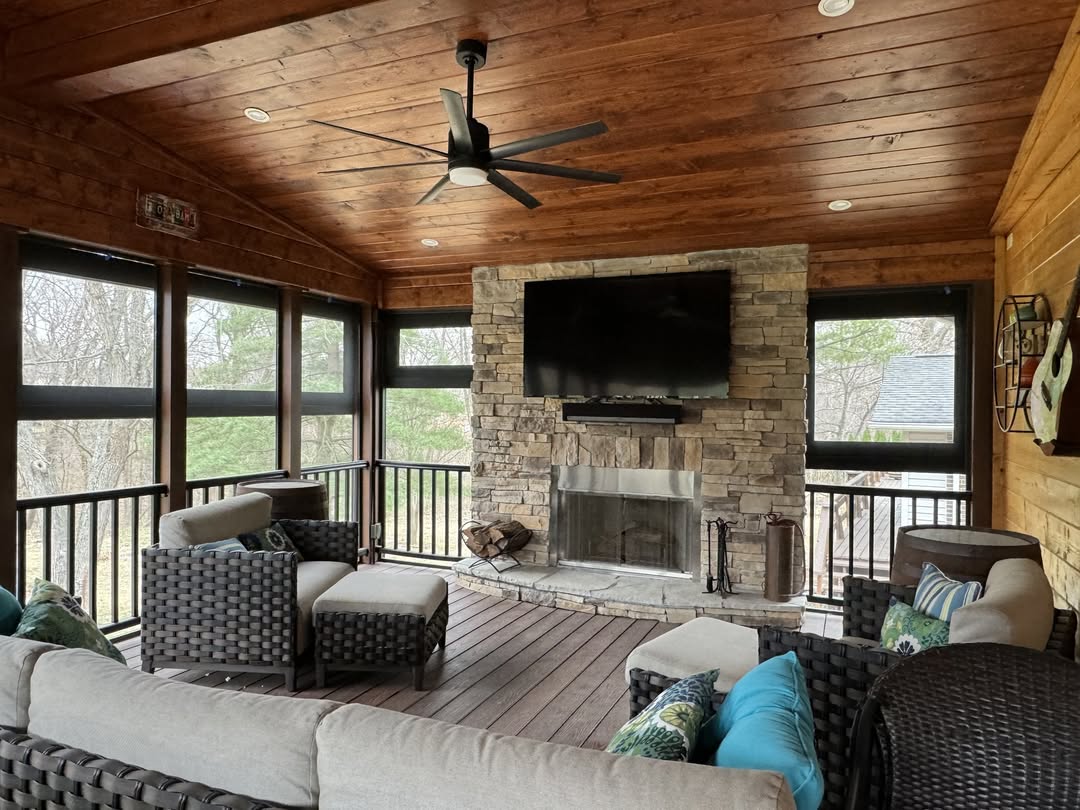 A cozy 3 season porch with wicker furniture, a stone fireplace, a mounted TV, and large windows overlooking trees—perfect for gathering new porch ideas.