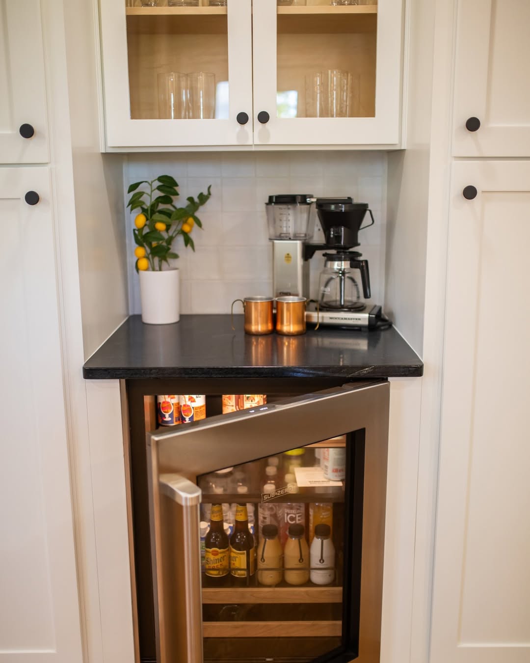A small beverage fridge with the door open shows drinks inside, below a black countertop—perfect for kitchen coffee bar ideas—with two copper mugs, a coffee maker, and a potted lemon plant. White cabinets surround the area.
