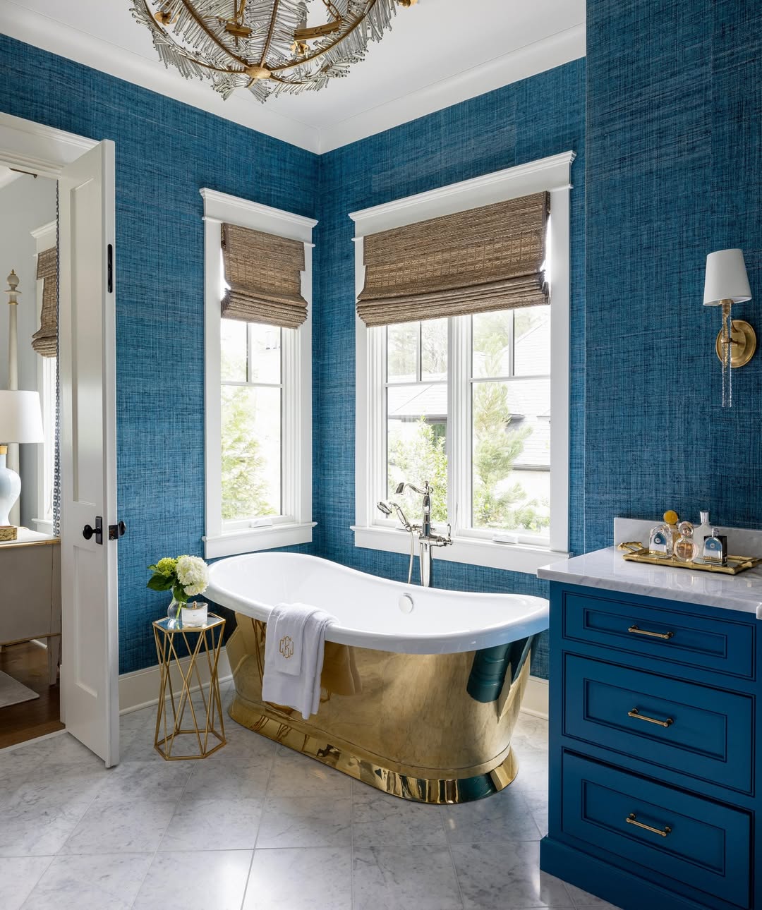 A bathroom showcasing inspiring tub design with blue textured walls, a gold freestanding bath, marble floor, blue vanity, and two windows with woven shades.