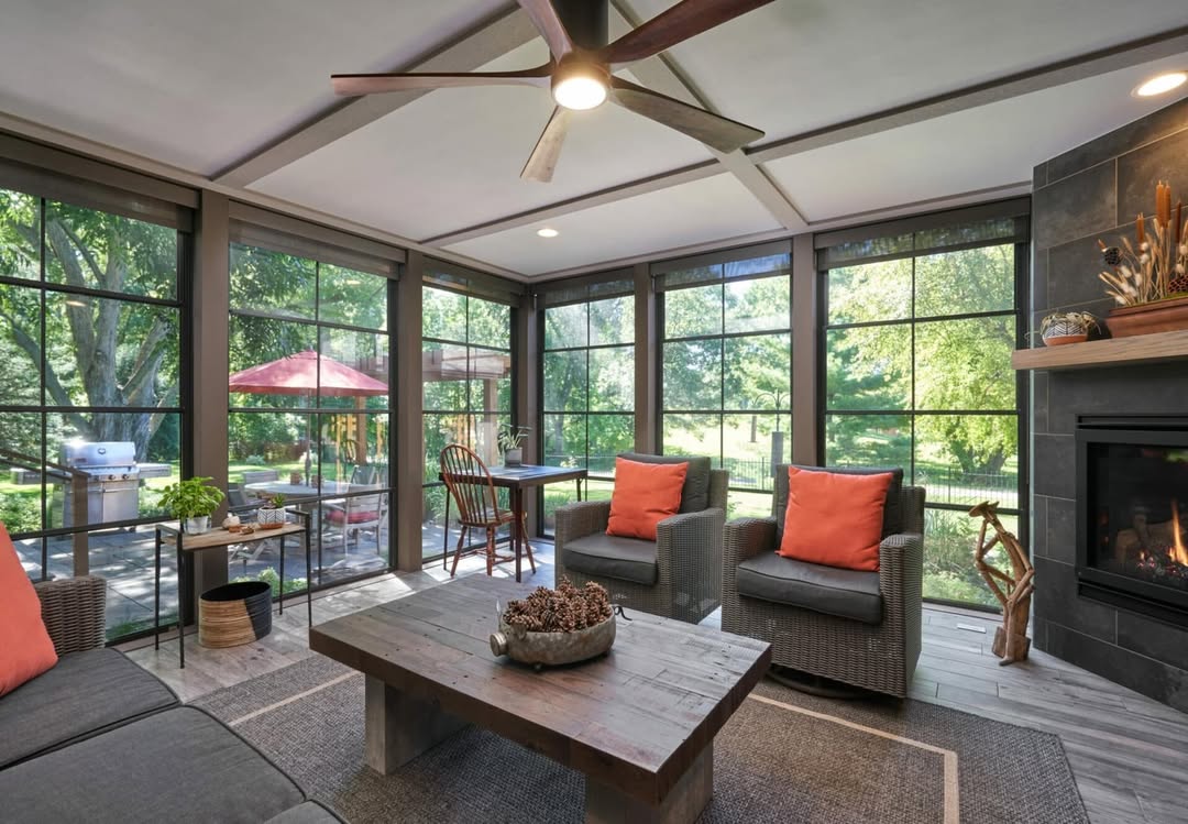 A modern 3 season porch with large windows, gray furniture with orange cushions, a wooden coffee table, fireplace, and a view of the patio and greenery outside—perfect for those seeking inspiring porch ideas.
