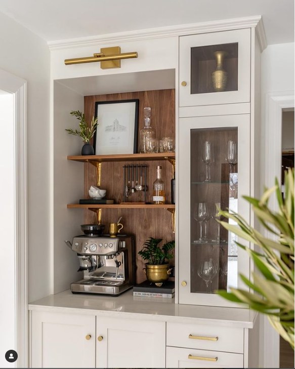 A built-in coffee bar with an espresso machine, glassware, gold accents, wooden shelves, decor items, and a plant in a white cabinet setting—perfect inspiration for your Kitchen Coffee Bar Ideas.