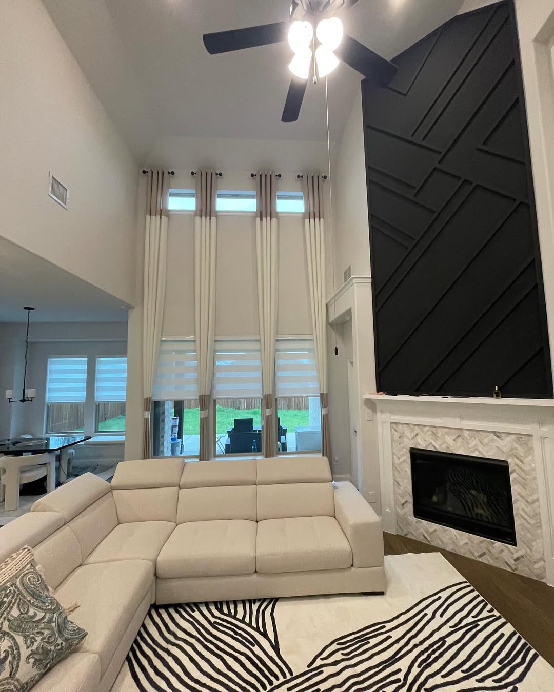 A modern high ceiling living room with tall windows and curtains, a black accent wall above a white fireplace, a beige sectional sofa, and a zebra-print rug—perfect inspiration for those seeking bigger home design ideas.