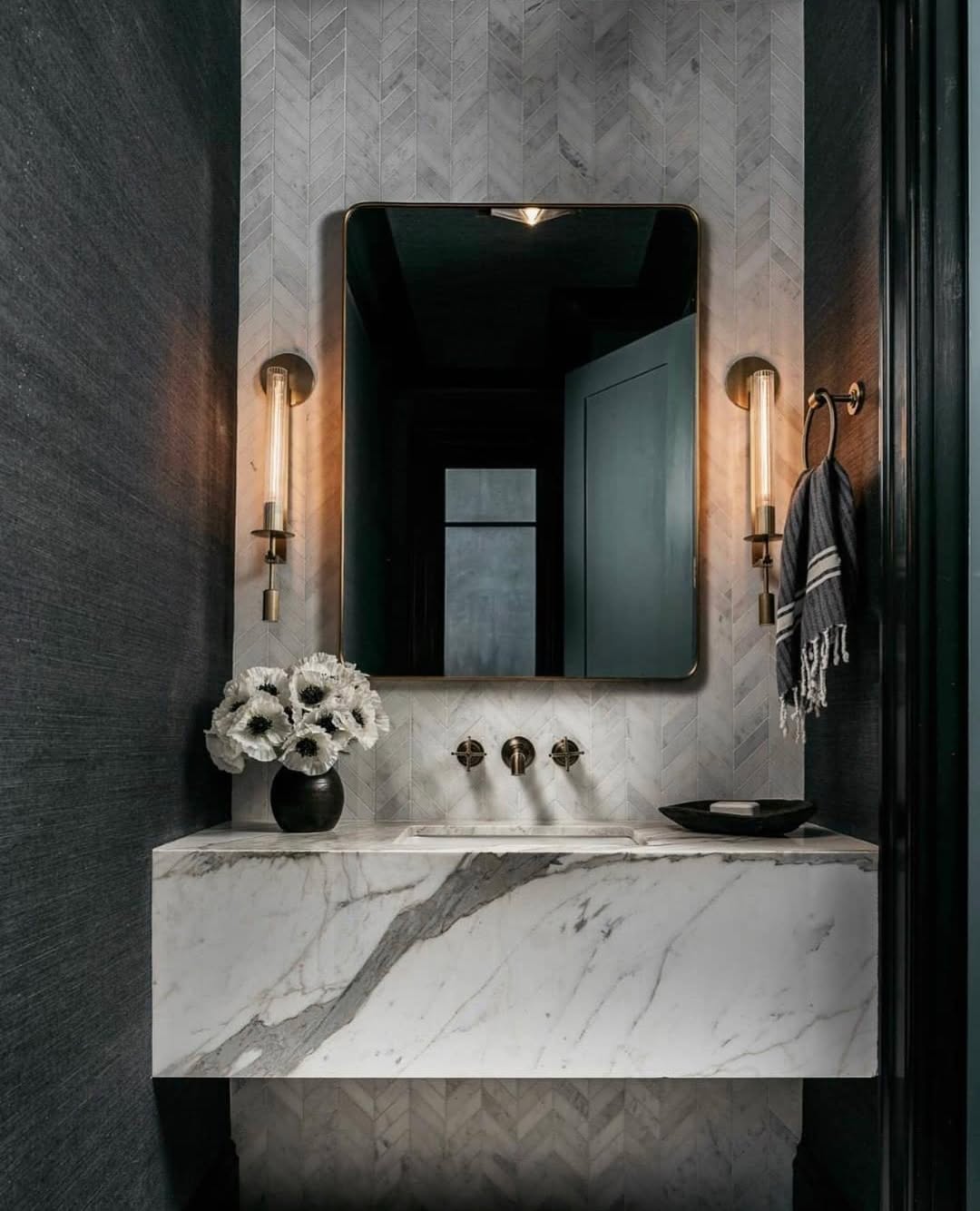Modern bathroom with a marble sink, black-framed mirror, wall sconces, towel ring with striped towel, and a vase of white flowers—perfect for anyone seeking stylish powder rooms or wow-factor bathrooms.
