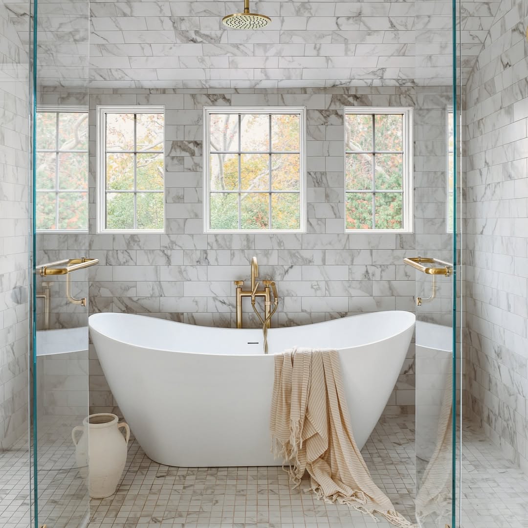 Modern bathroom with a freestanding white bathtub, gold fixtures, a towel draped over the tub, marble tile walls, three windows, and a ceramic jug on the floor—perfect for those seeking Bathroom Tub Ideas.