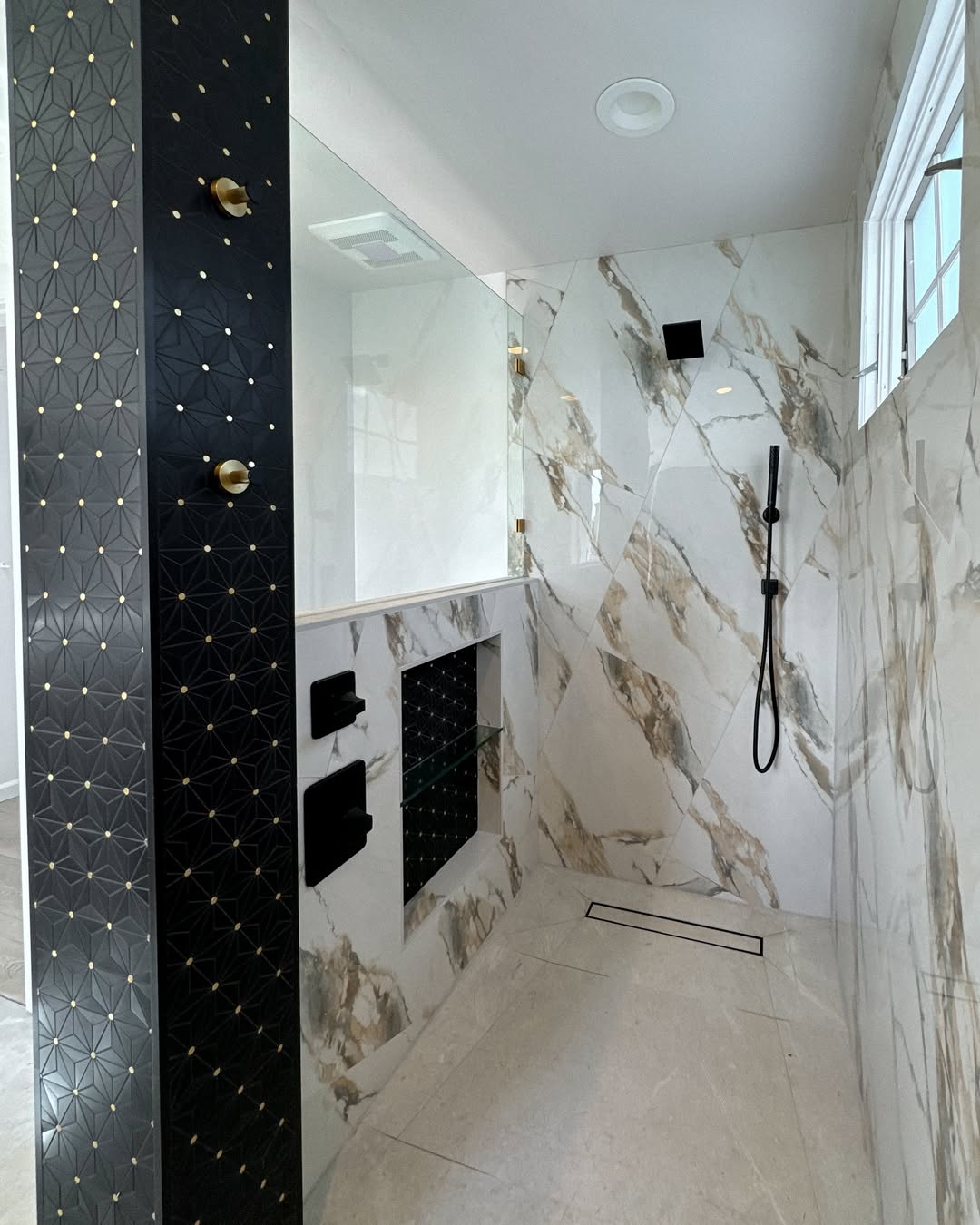 Modern walk-in shower with white and gold marble walls, black fixtures, a geometric black accent wall, a handheld showerhead, and a linear floor drain—perfect for no door shower designs and small bathroom ideas.