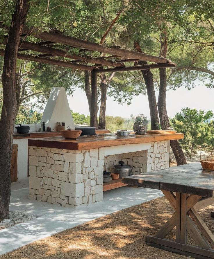 Rustic outdoor kitchen featuring a stone counter, wooden pergola, and wooden dining table set beneath trees, with sunlight and greenery providing the perfect backdrop for inspired outdoor kitchen ideas.