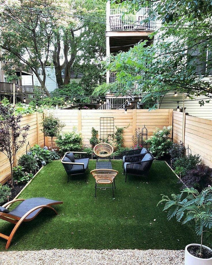 A small backyard with artificial grass, modern lounge chairs, a wicker patio set, and wooden fencing, surrounded by plants and trees—perfect for those seeking inspiring small backyard landscaping ideas.