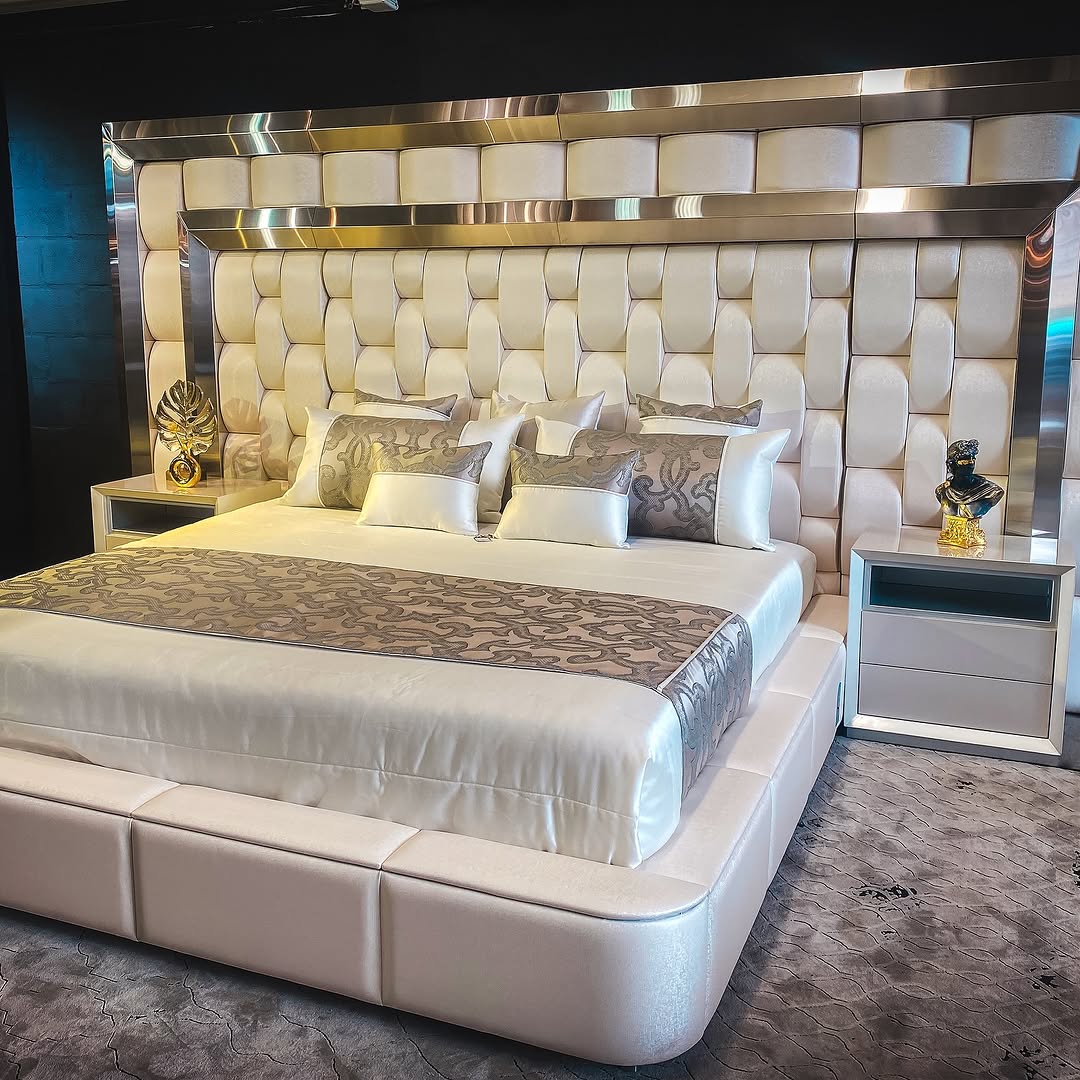 A modern Italian bedroom with a large upholstered bed, patterned pillows and bedding, two white nightstands, and metallic decorative accents.
