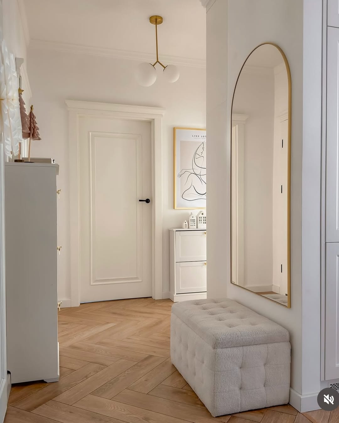 A bright small entryway featuring a herringbone wooden floor, white tufted bench, tall arched mirror, small dresser, framed art print, and a white door with a black handle.