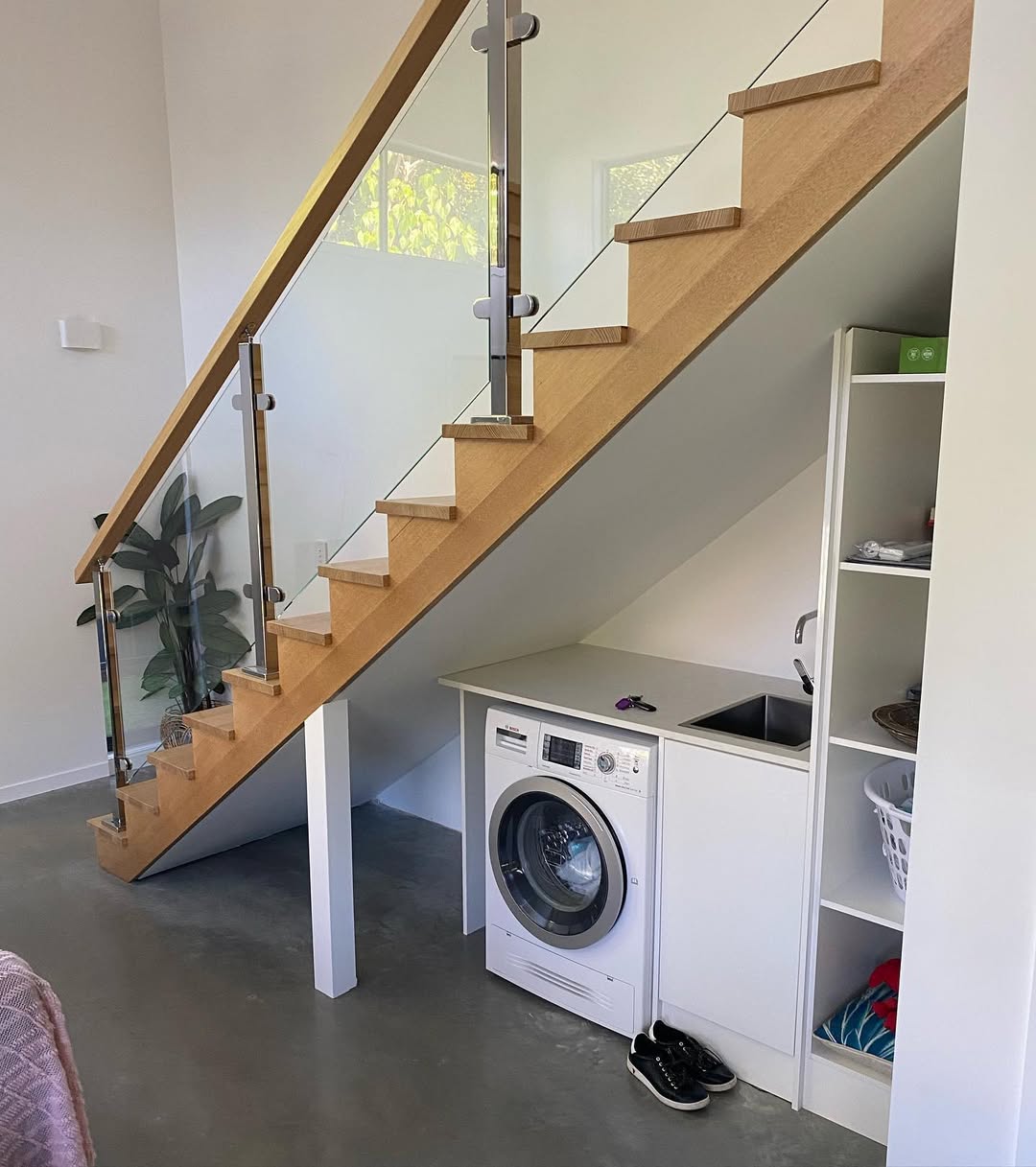 An under stair laundry room features a washing machine, sink, and open shelving installed beneath a wooden staircase with a glass railing in a modern, minimalistic interior.