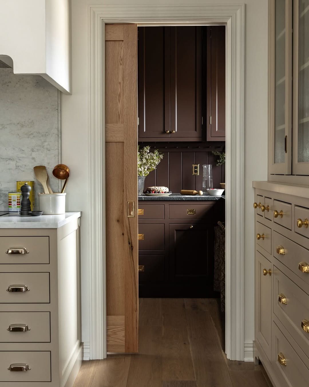 A partially open wooden sliding door adds a wow factor, separating a light-toned kitchen with brass hardware from a darker pantry featuring brown cabinets and decorative items on the counter—perfect for real life pantry door ideas.