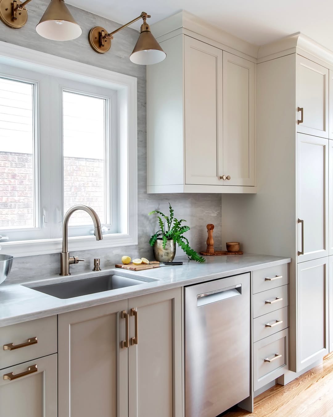 A modern galley kitchen design featuring light beige cabinets, a stainless steel dishwasher, single-basin sink, brushed metal fixtures, and a potted plant by the window.