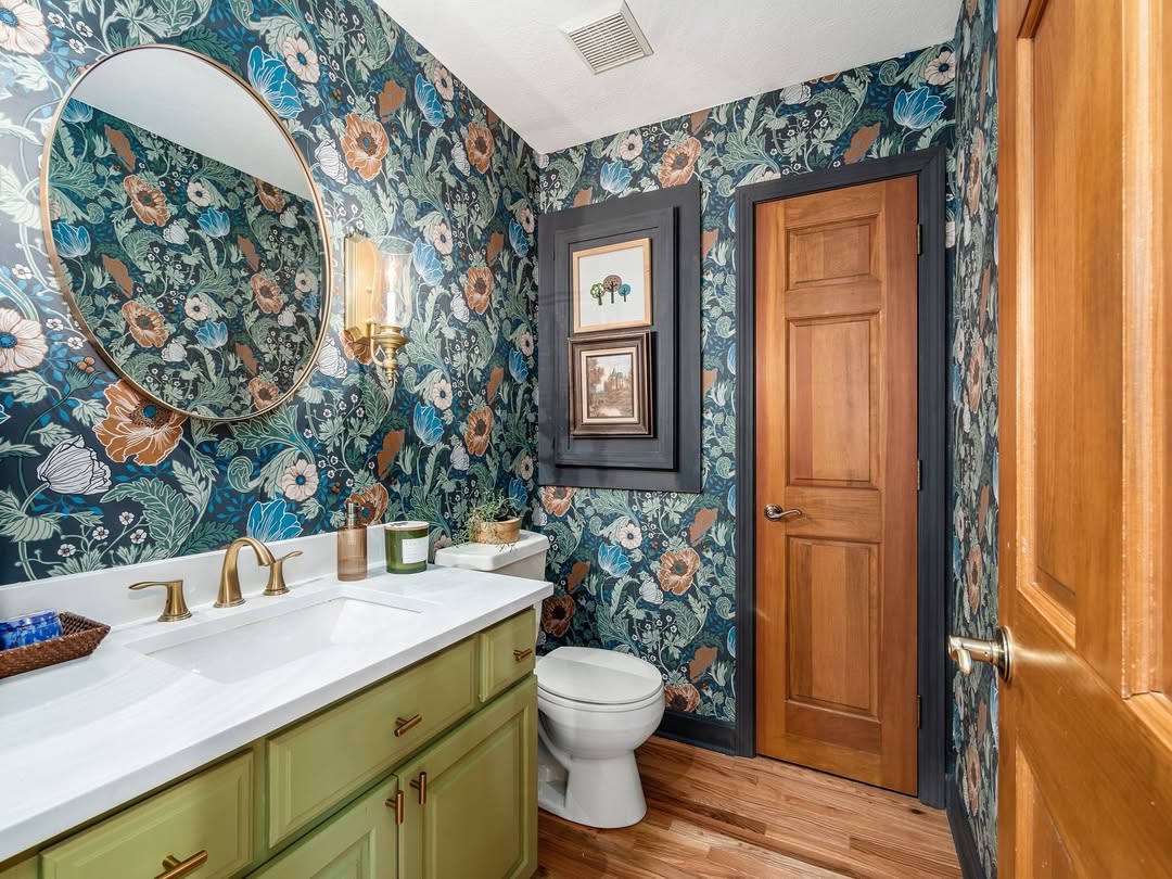 This bathroom features wow-factor decor with floral wallpaper, a green vanity with gold fixtures, a white countertop, round mirror, wooden door, and framed art above the toilet—perfect for stylish powder room ideas.