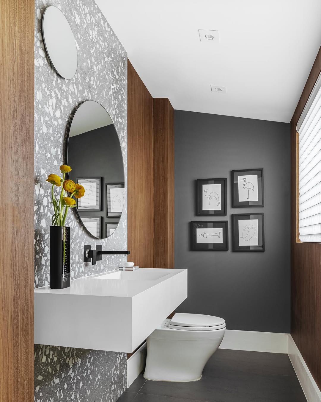 Modern bathroom with wood panel walls, terrazzo accent behind a round mirror, white floating sink, vase with yellow flowers, toilet, and black-and-white framed artwork—perfect inspiration for stylish powder rooms seeking wow-factor decor.