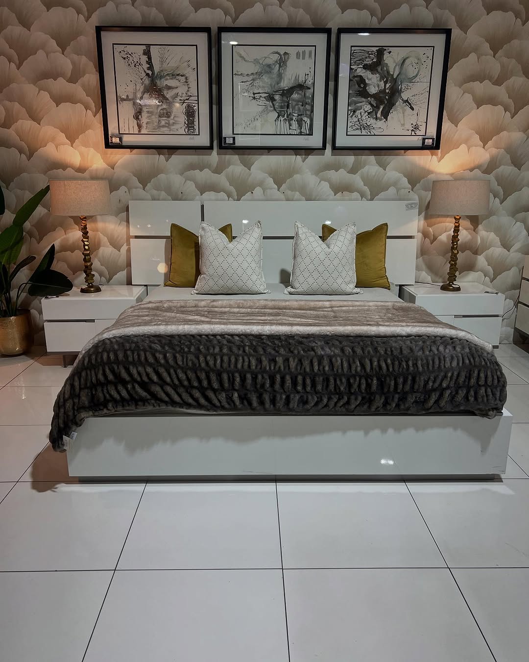 Modern Italian bedroom with a white bed, gray throw, gold and white pillows, two bedside tables with lamps, and three abstract paintings on a floral-patterned wall.