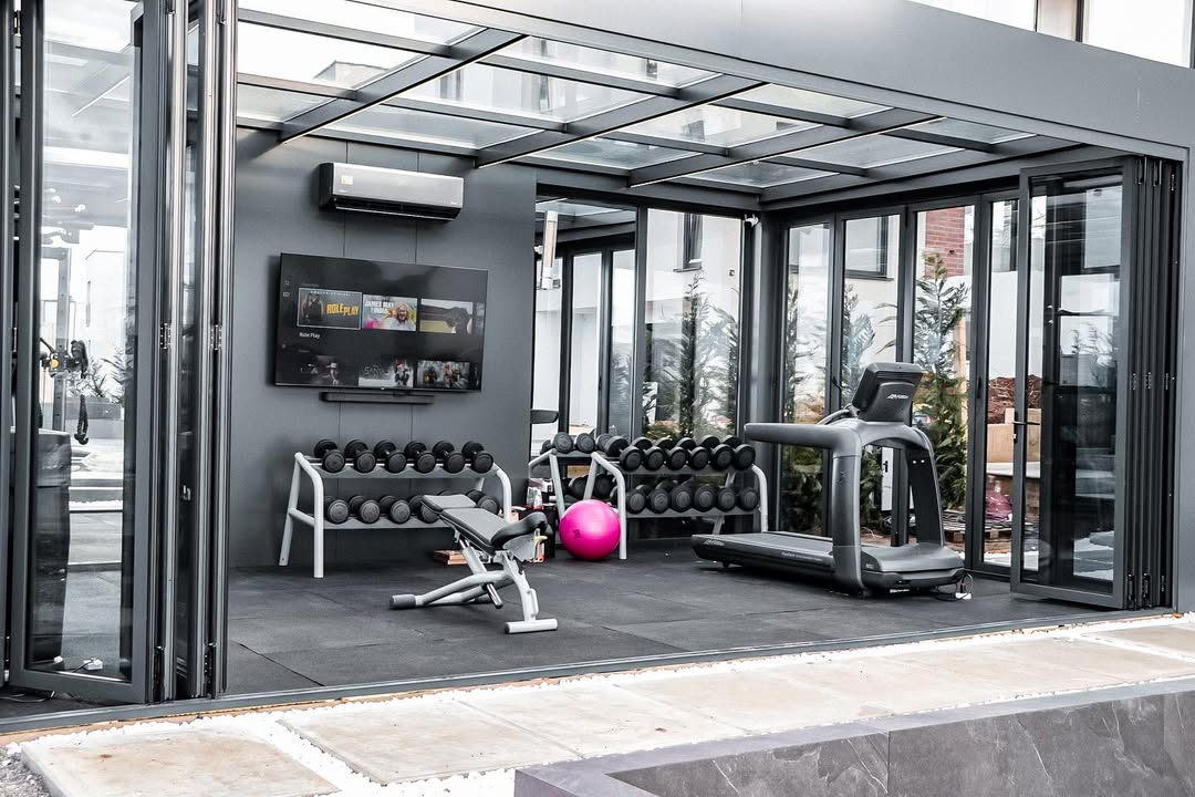 A stylish mini gym at home featuring a treadmill, weight bench, dumbbells, wall-mounted TV, pink exercise ball, and sleek glass walls.