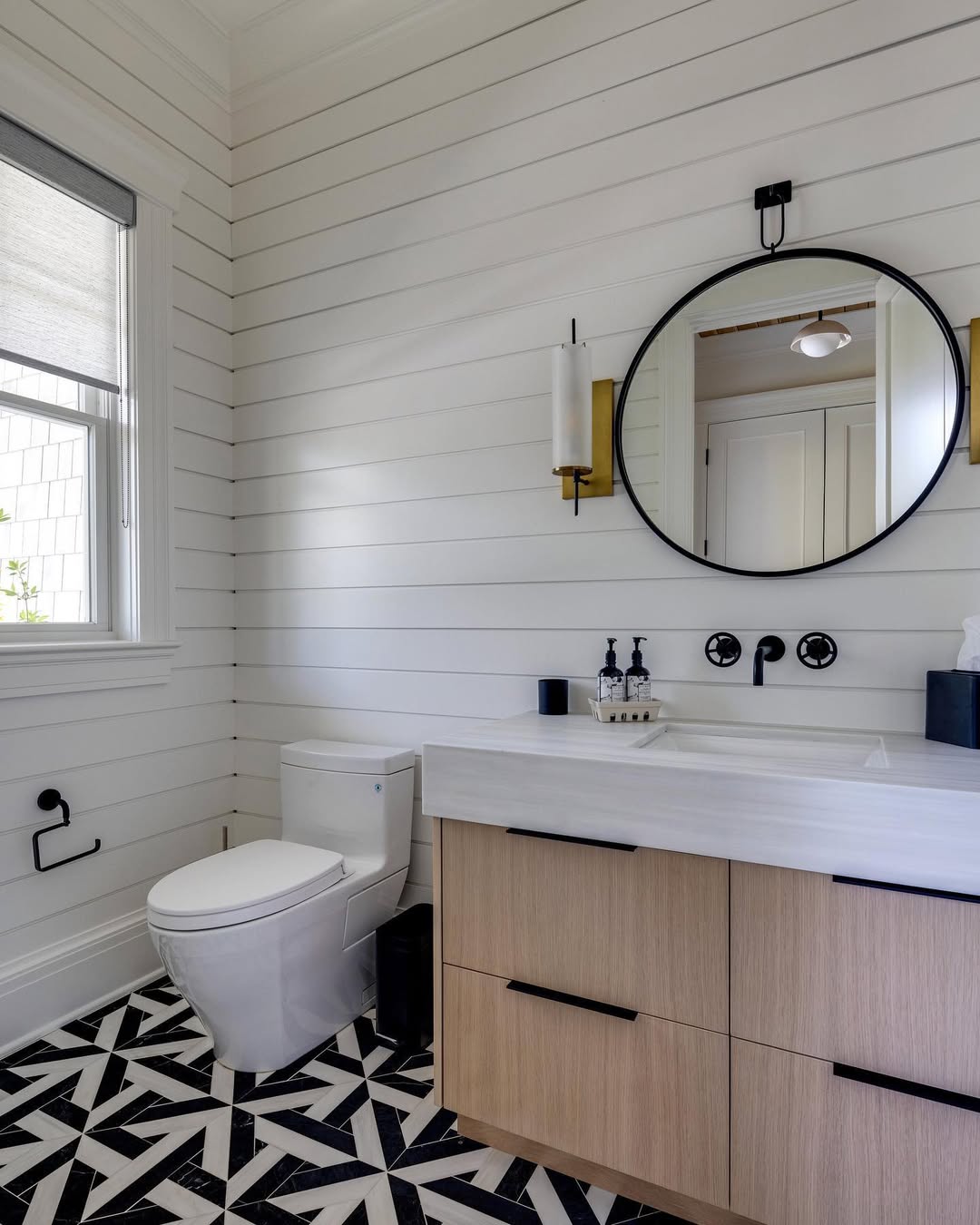Modern shiplap wall bathroom with white shiplap walls, black and white geometric tile floor, a round mirror, wall-mounted faucet, light wood vanity, and a window with a roller shade.