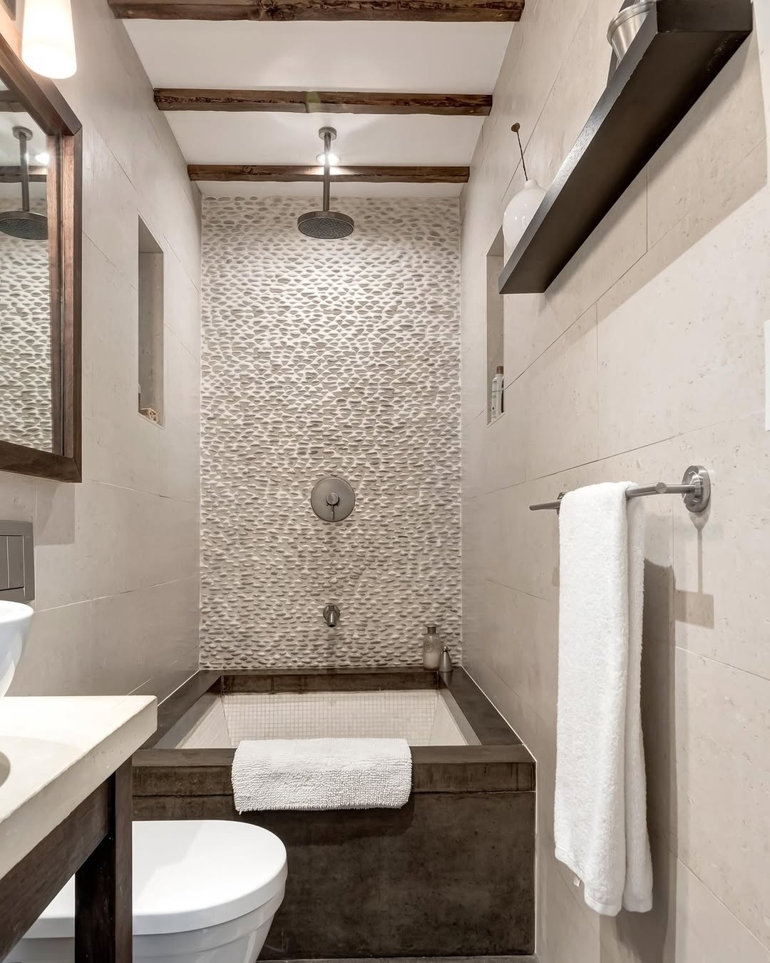 Narrow small bathroom with a modern walk-in shower and bathtub, mosaic tile accent wall, exposed wooden ceiling beams, and white towels on a rail and tub.