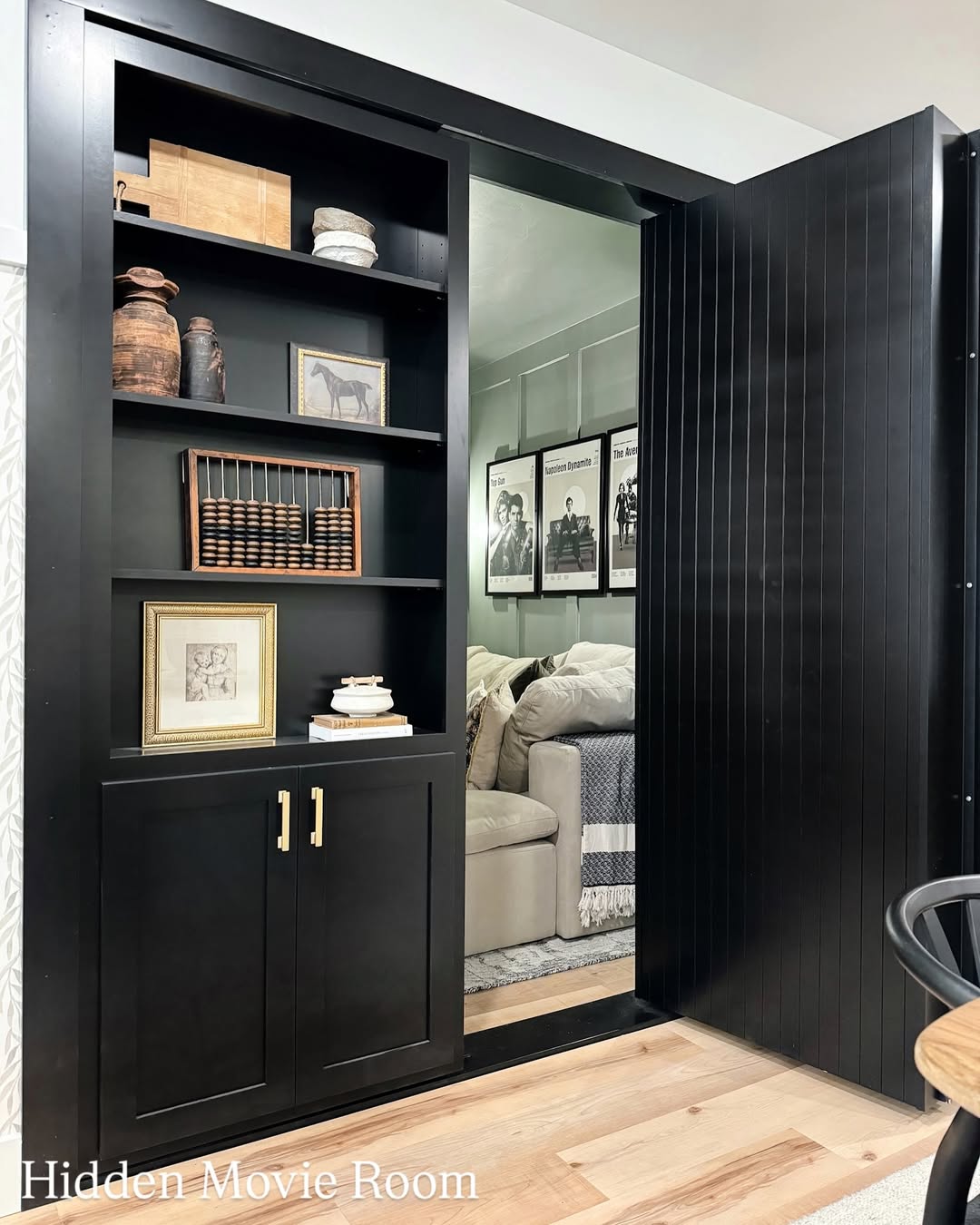 A black built-in bookshelf with a sliding door partially open, revealing a hidden movie room with sofas and framed art—perfect inspiration for stylish homeowners seeking unique hidden door ideas.