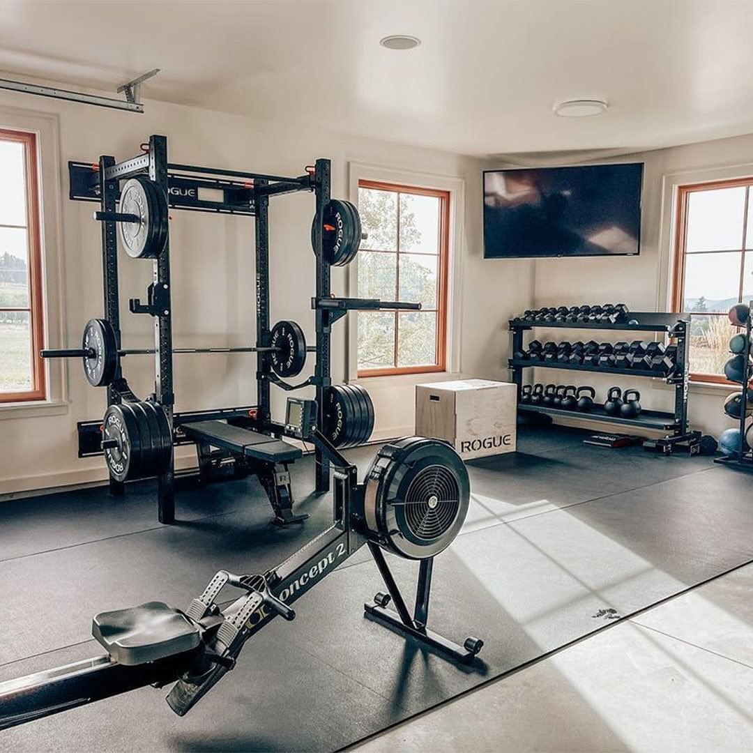 Create your ideal mini gym at home with a rowing machine, squat rack, weight plates, dumbbells, medicine balls, a plyo box, and a wall-mounted TV—all in a bright room with large windows.