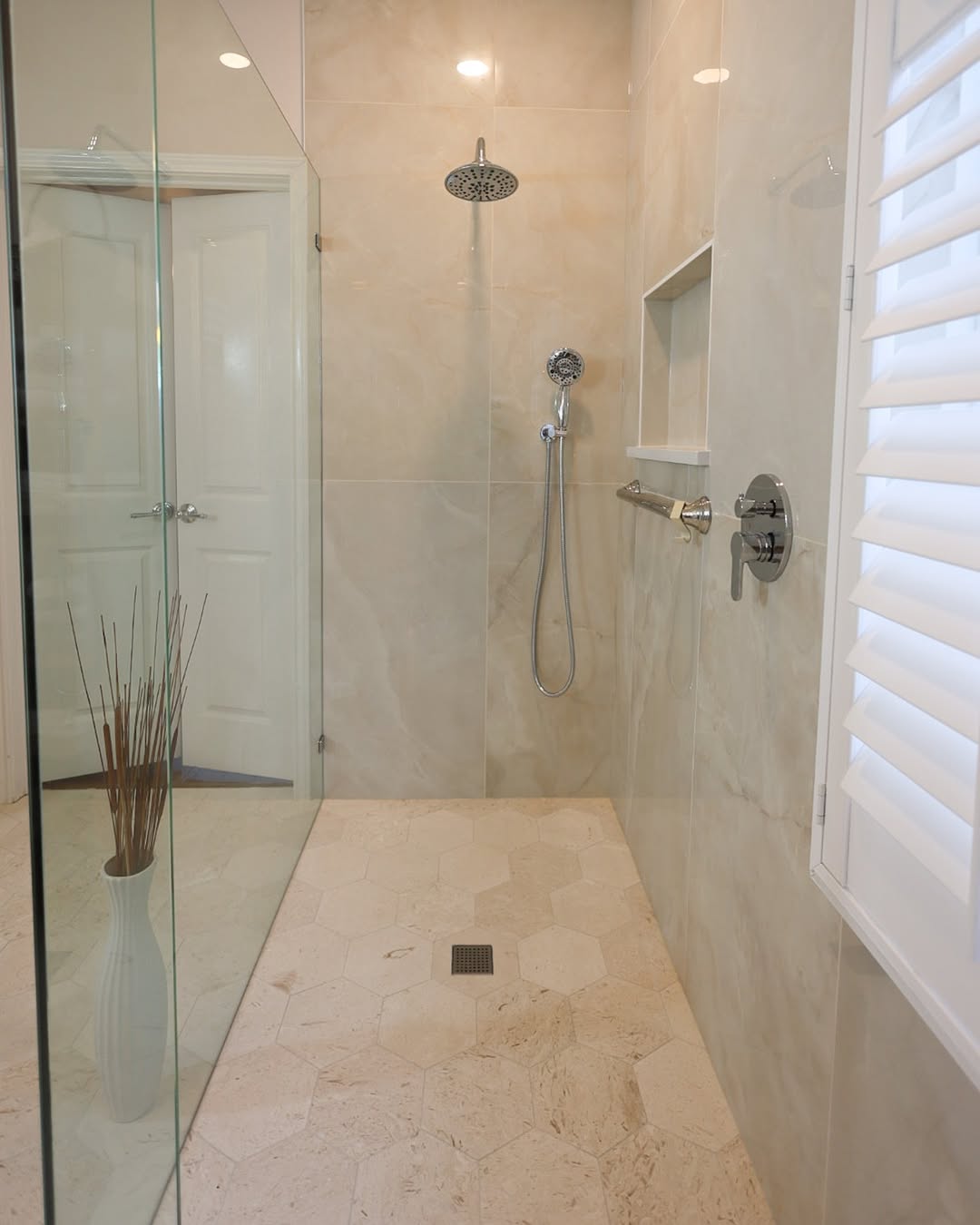 Modern walk-in shower with beige hexagonal floor tiles, wall-mounted showerhead, handheld shower, grab bar, built-in shelf, and glass enclosure—perfect inspiration for walk-in shower ideas in small bathrooms. White shutters and a decorative vase are visible.