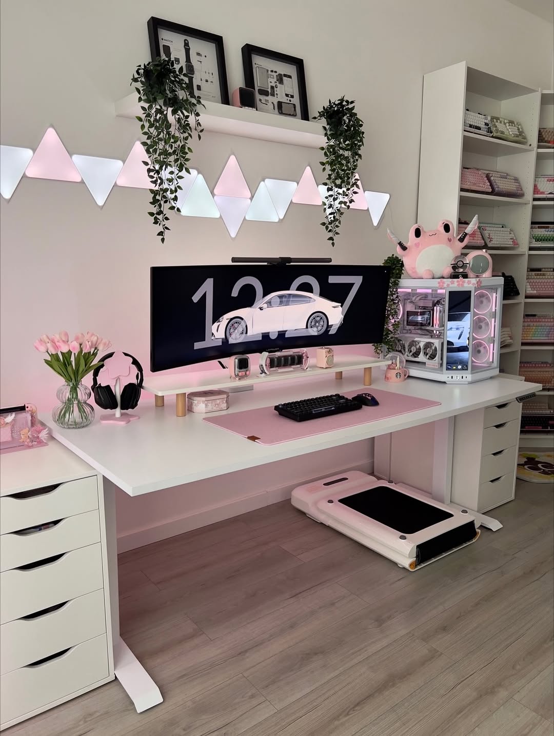 A modern computer desk setup with a large monitor, white accessories, pink lighting, hanging plants, and organized shelves in a bright room—perfect for anyone seeking desk organization ideas.