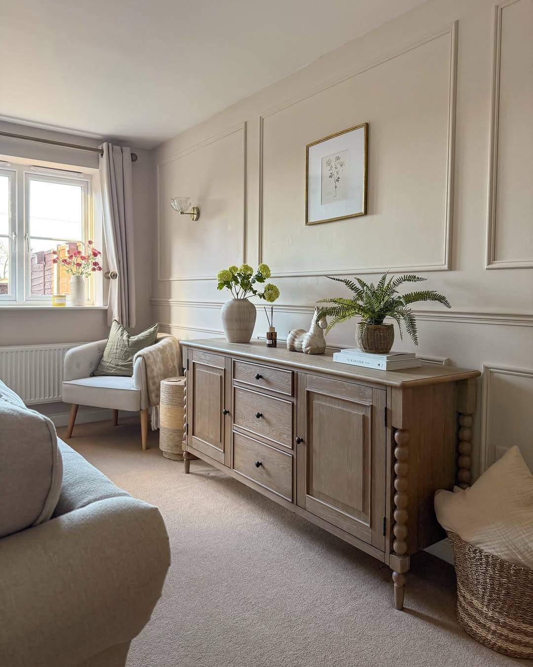 A beige living room with a wooden sideboard featuring tasteful sideboard decoration, potted plants, a framed botanical print, an armchair by the window, and neutral decor elements.
