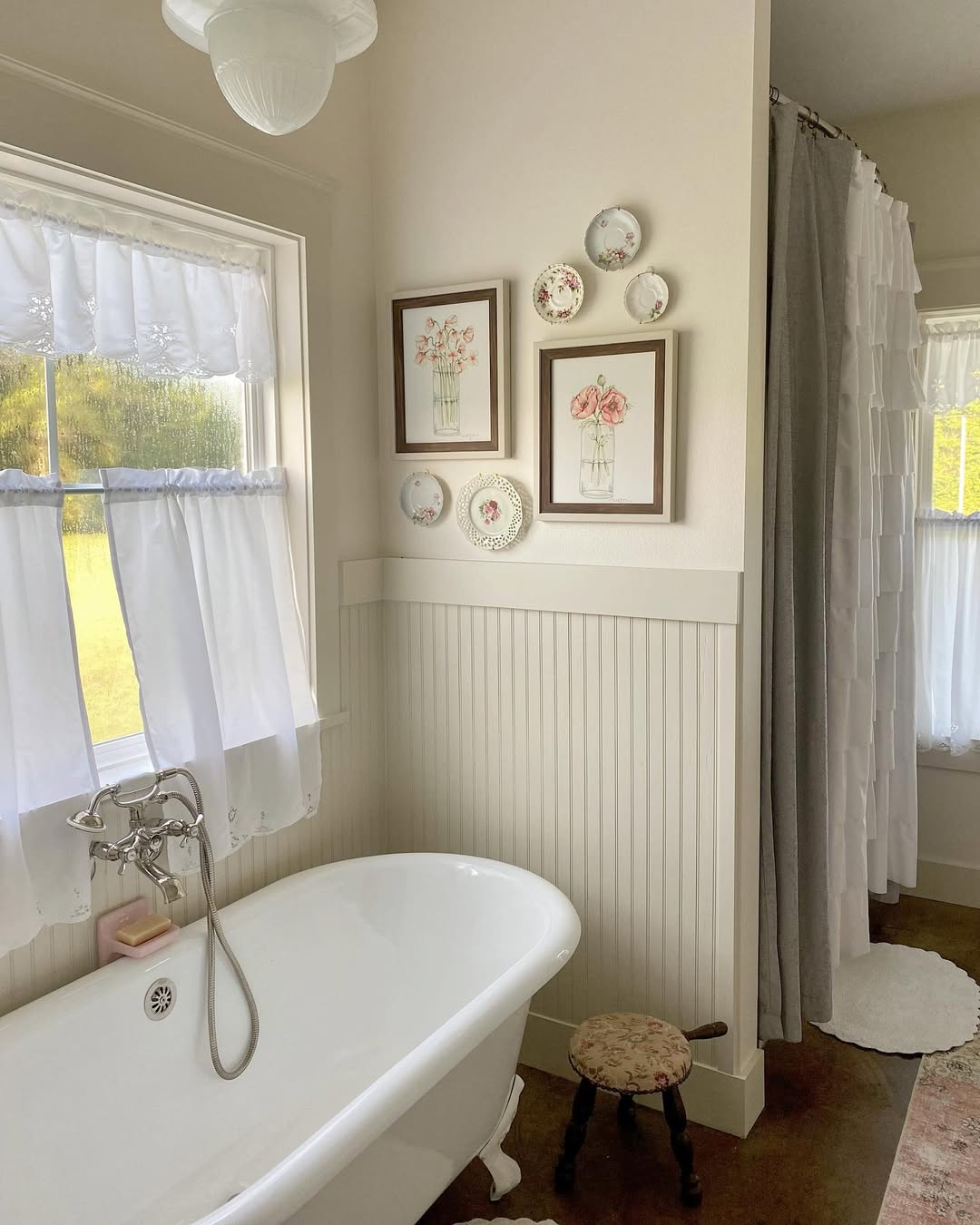 A bathroom featuring a white clawfoot tub showcases timeless tub designs, with a half-curtained window, framed floral art, decorative plates on the wall, a small stool, and a shower with a ruffled white curtain.