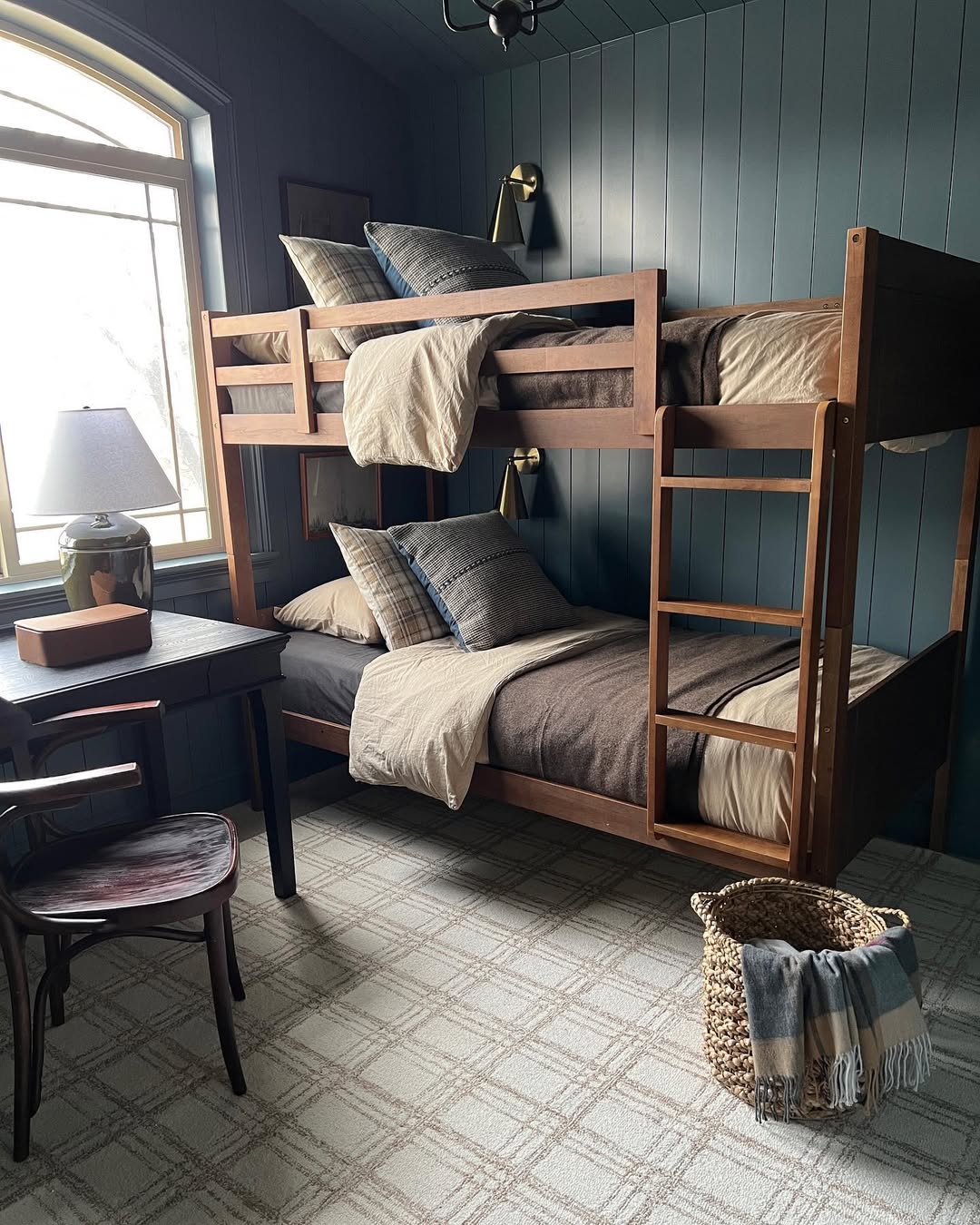 A tidy boys bedroom featuring wooden bunk beds, neutral bedding, a desk with a lamp, chair, and clever storage solutions like a woven basket with a blanket on a patterned carpet.