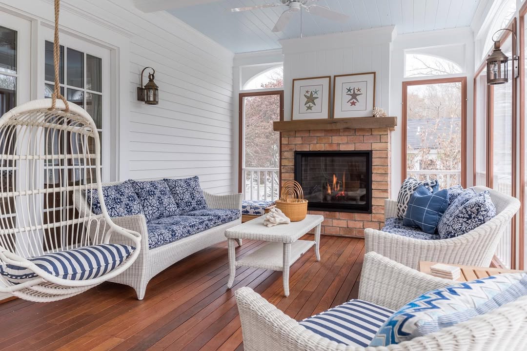 A sunroom with wicker furniture, blue and white cushions, a hanging chair, a wooden coffee table, and a brick fireplace with framed art above it offers inviting 3 season porch ideas for stylish porch decorating.