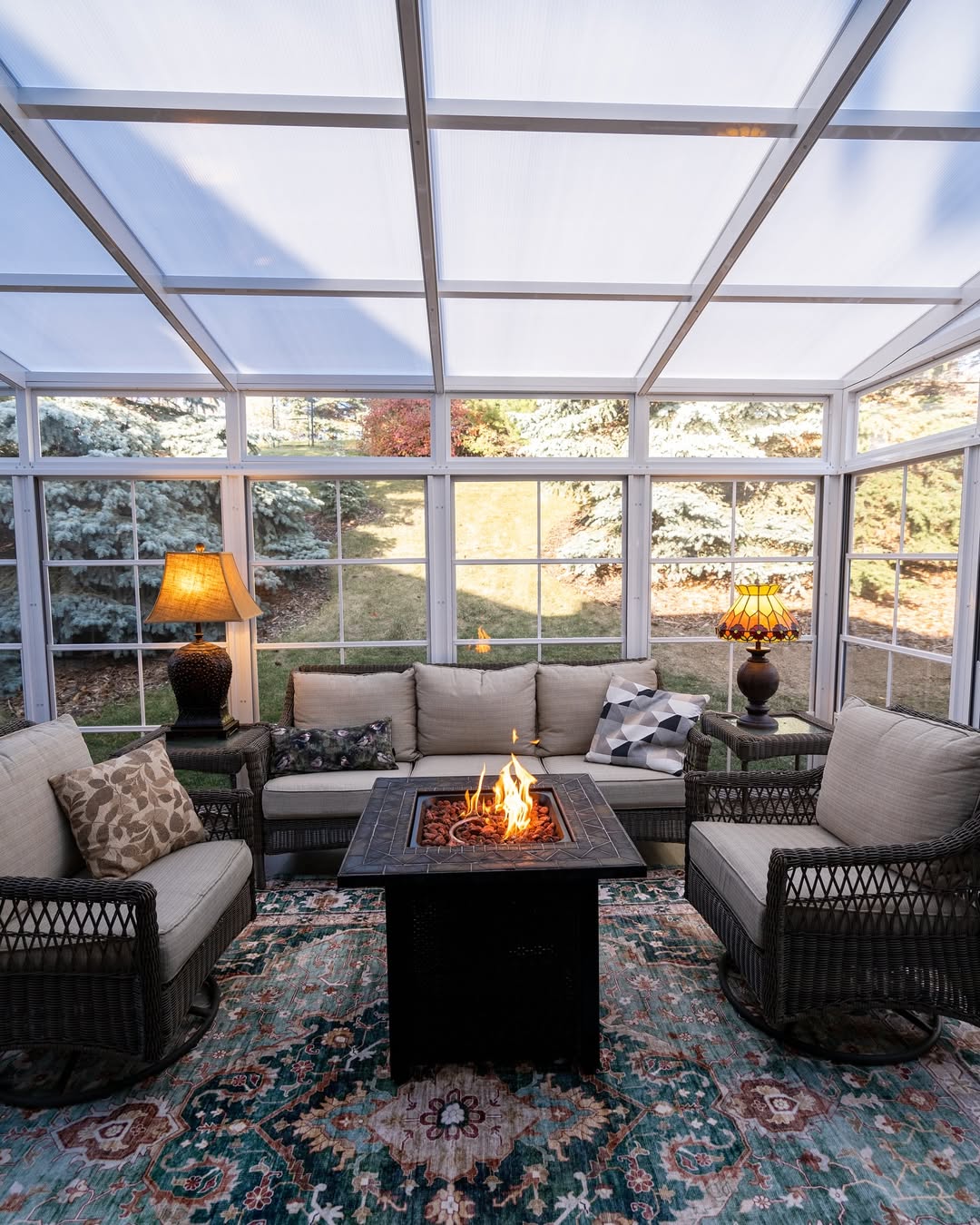 A sunroom designed as a 3 season porch, featuring wicker furniture, a patterned rug, two lamps, and a fire pit table, surrounded by large windows overlooking the yard—perfect for inspiring your next porch ideas.