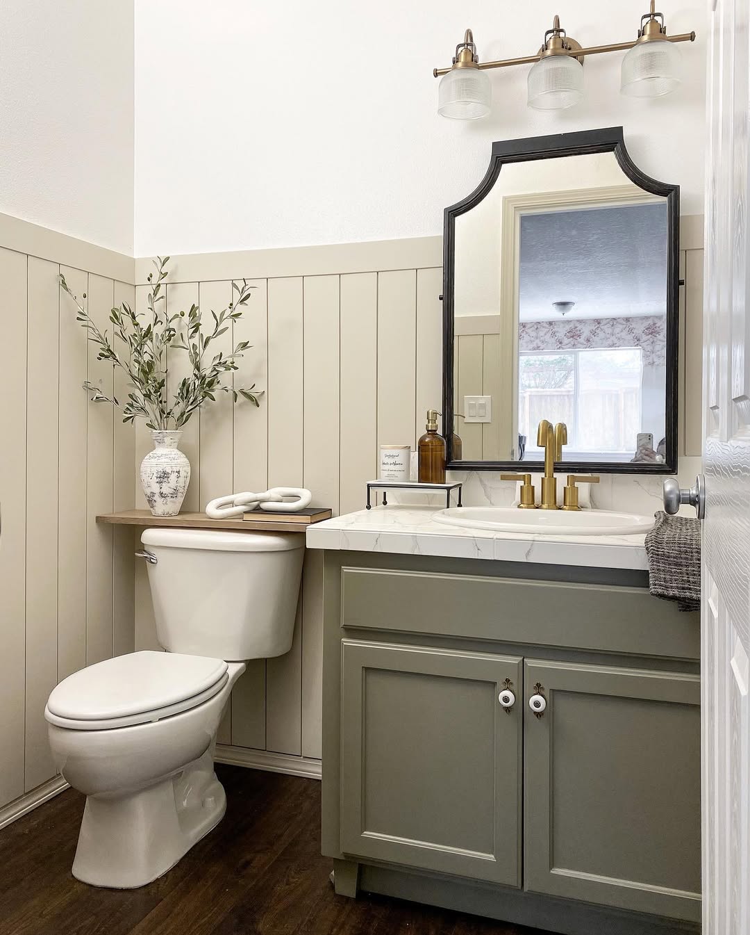 Modern shiplap wall bathroom featuring a green vanity, marble countertop, round mirror, white toilet, potted plant, and brass light fixture on wood-paneled walls.