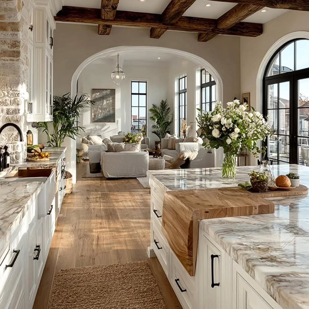 A spacious kitchen with marble countertops and wood accents showcases modern kitchen ideas, opening to a bright living area with large windows, white sofas, and abundant natural light.
