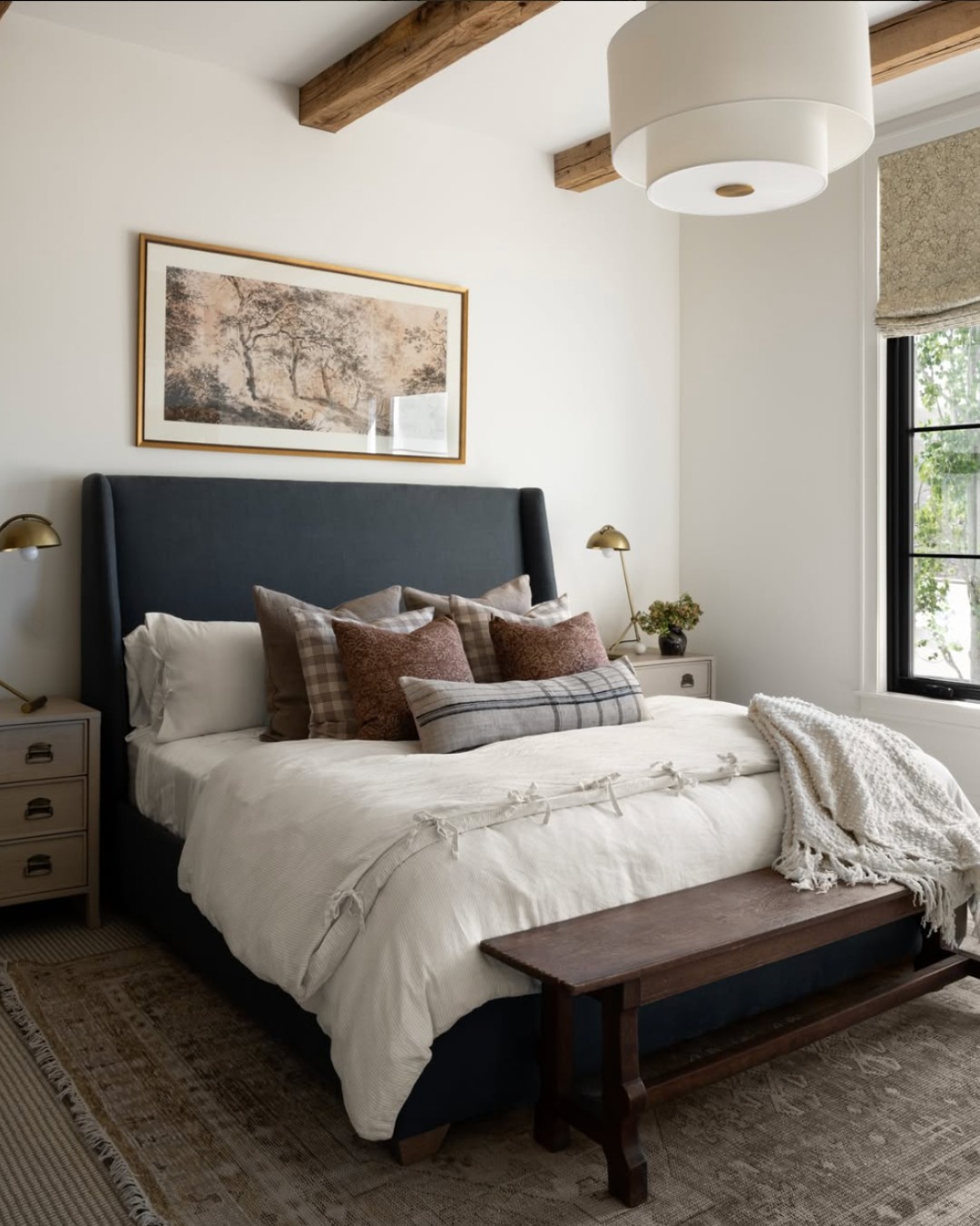A neatly made bed with neutral bedding, plaid pillows, and a throw sits in a cozy guest bedroom featuring wood ceiling beams, a large window, and framed tree artwork above the headboard—perfect for inspiring guest bedroom ideas.