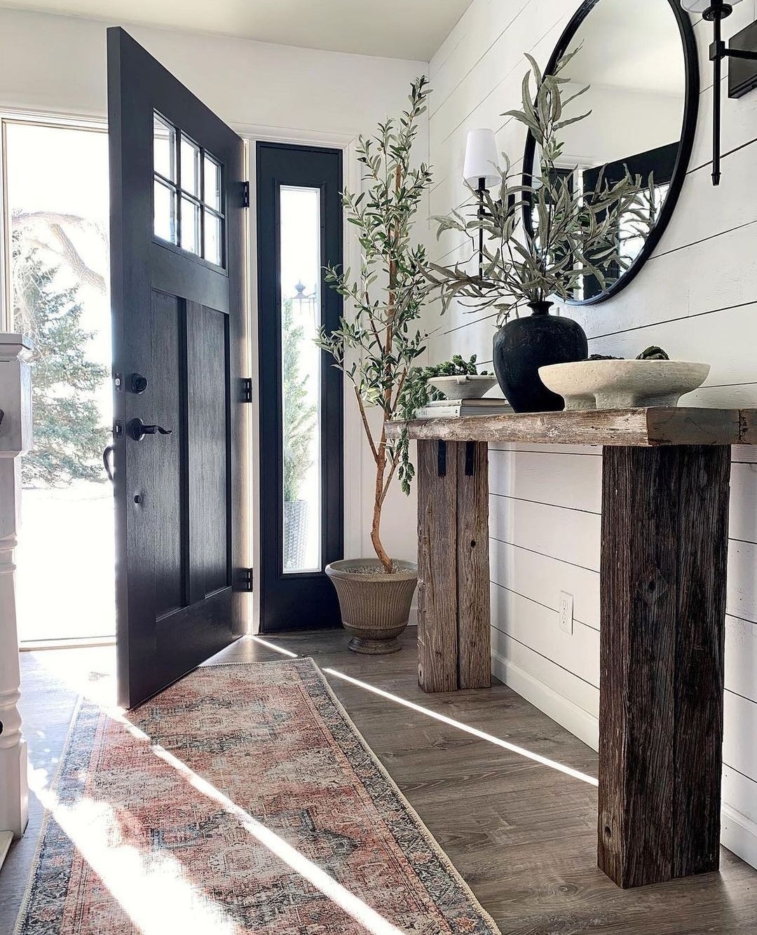 A rustic entryway features a wooden console table with vases, a round mirror, a potted plant, and a patterned rug on wood flooring next to an open black door—perfect inspiration for modern entryway ideas.
