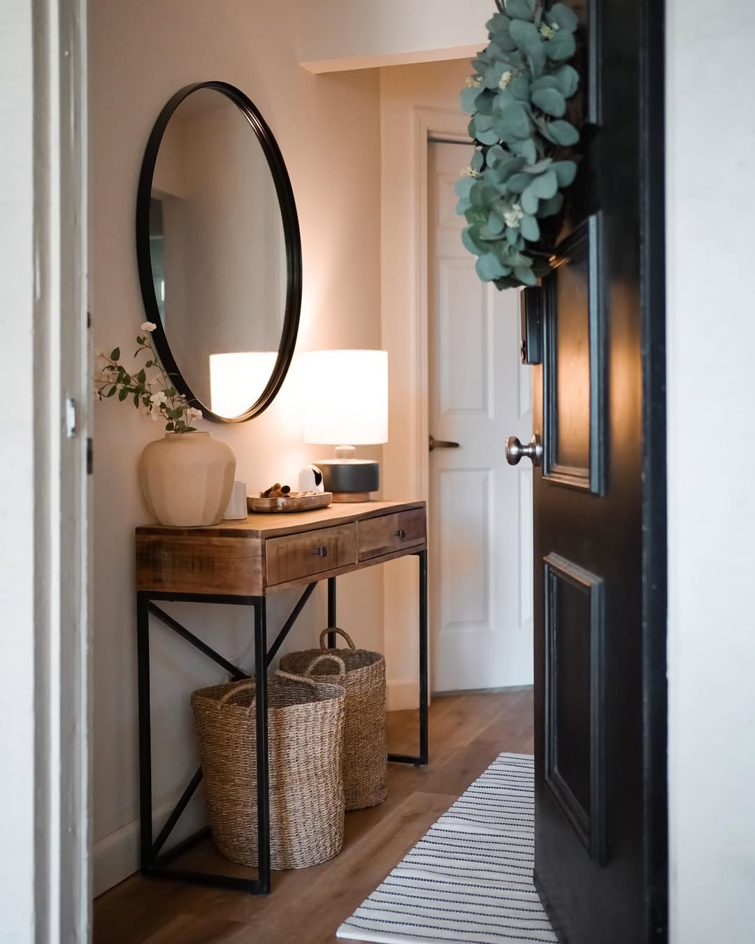 A tidy small entryway with a black door, a wooden console table, a round mirror, a lamp, decorative vase, and two woven baskets underneath.